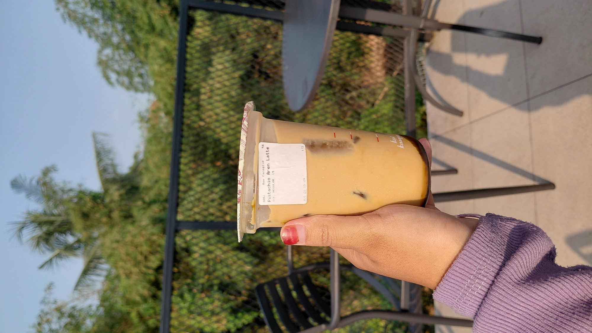 A hand with a red-painted fingernail holds a plastic cup of iced coffee. The cup has a white label that reads "Pistachio Aren Latte" and "AUG 21 15:24". The drink is a pale brown color with ice cubes visible. The background is blurred and shows greenery, a metal fence, a black chair, and a round table. Sunlight casts shadows on the tiled ground. The scene appears to be outdoors at a cafe.