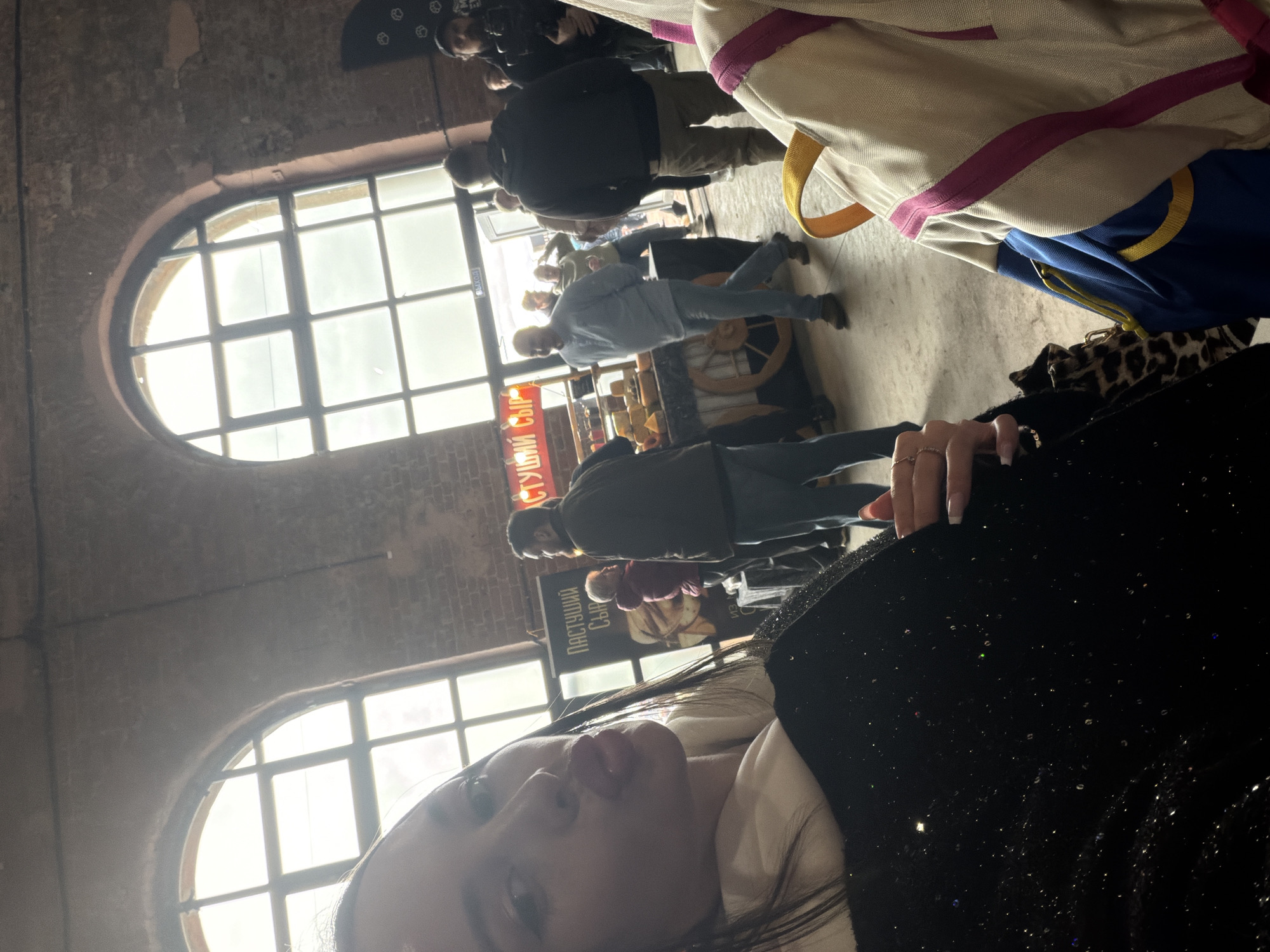 The image, taken indoors, shows a woman in the foreground with long dark hair, light skin, and full lips. She is wearing a white top with a black, glittery garment. Her hand is visible with rings on her fingers.
In the background, there's a crowded indoor market or fair. People are milling around stands and displays. Arched windows with multiple panes are visible in the background, admitting bright daylight. The walls of the building appear to be made of brick. A sign in Cyrillic script can be seen behind the crowd.
Given the architecture and the script, the location is likely in Saint Petersburg, Russia.