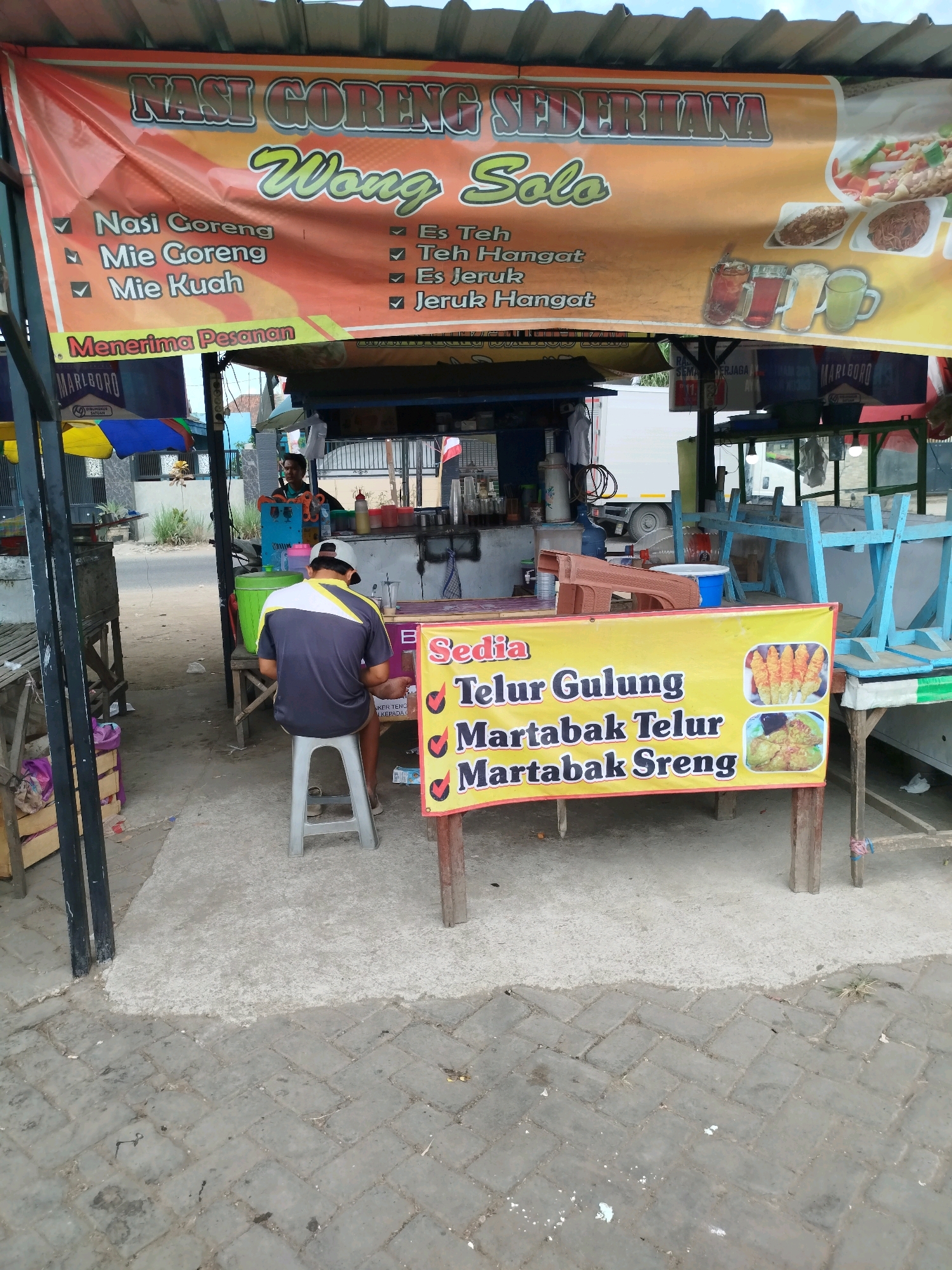 The image depicts an outdoor food stall in Burneh, Indonesia. A large banner with the text "Nasi Goreng Sederhana Wong Solo" and a list of food and drink items such as "Nasi Goreng", "Mie Goreng", "Mie Kuah", "Es Teh", "Teh Hangat", "Es Jeruk", and "Jeruk Hangat" is prominently displayed. Below this, another yellow banner advertises "Sedia Telur Gulung, Martabak Telur, Martabak Sreng" with accompanying images of these food items.
A person is seated on a stool in front of the stall, wearing a grey and yellow shirt and a grey cap. Another person is visible standing behind the counter of the stall, appearing to be working. The stall is set up on a concrete and paved ground area. Various items associated with a food stall, such as cooking equipment, containers, and furniture, are present. In the background, a truck and some buildings can be seen. The overall scene suggests a bustling street food environment.