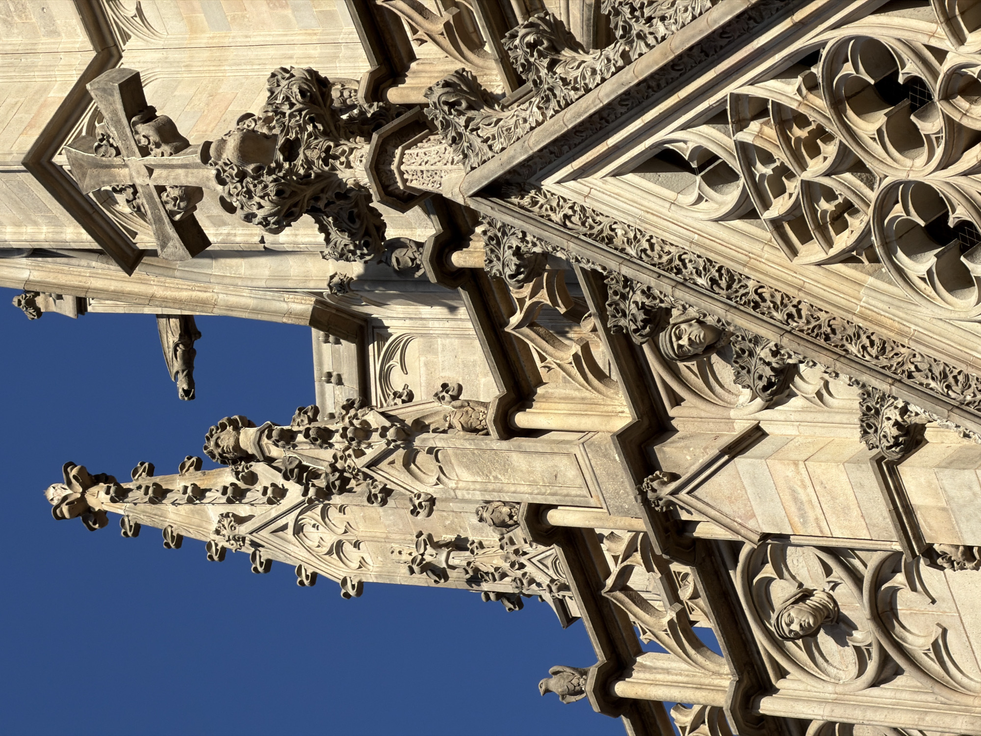 This image captures a close-up, angled view of intricate Gothic architecture, likely from a cathedral or historical building, under a clear, bright blue sky. The sun is casting strong shadows, suggesting it is midday. The stone carvings are highly detailed, featuring ornate floral motifs, geometric patterns, and what appear to be gargoyles or grotesque figures. One prominent detail is a carved cross with small, human-like faces incorporated into its design. Further along the structure, a series of circular openings with elaborate tracery are visible, and one of these openings contains a carved human face looking outwards. The architectural elements suggest a grand and ancient edifice, indicative of European Gothic style. The overall impression is one of historical grandeur and meticulous craftsmanship.