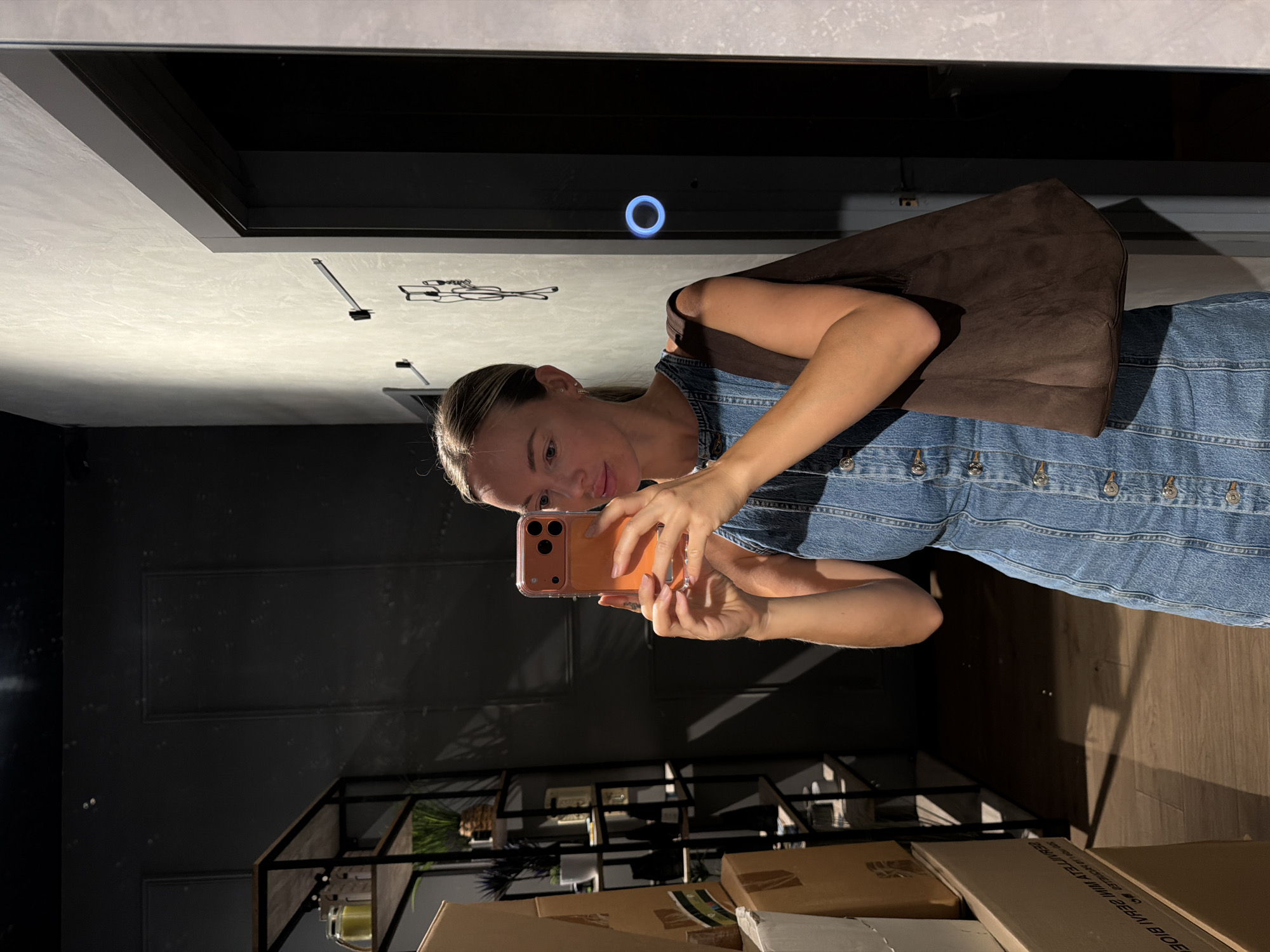 In a modern, dimly lit interior, a woman with blonde hair pulled back is taking a selfie in a mirror. She is wearing a denim dress and carrying a brown tote bag over her shoulder. The background features shelves with various items, cardboard boxes, and a doorway with a blue illuminated ring. The overall ambiance suggests a personal space, possibly a dressing room or a stylish shop, located in the city of Benidorm, Spain.