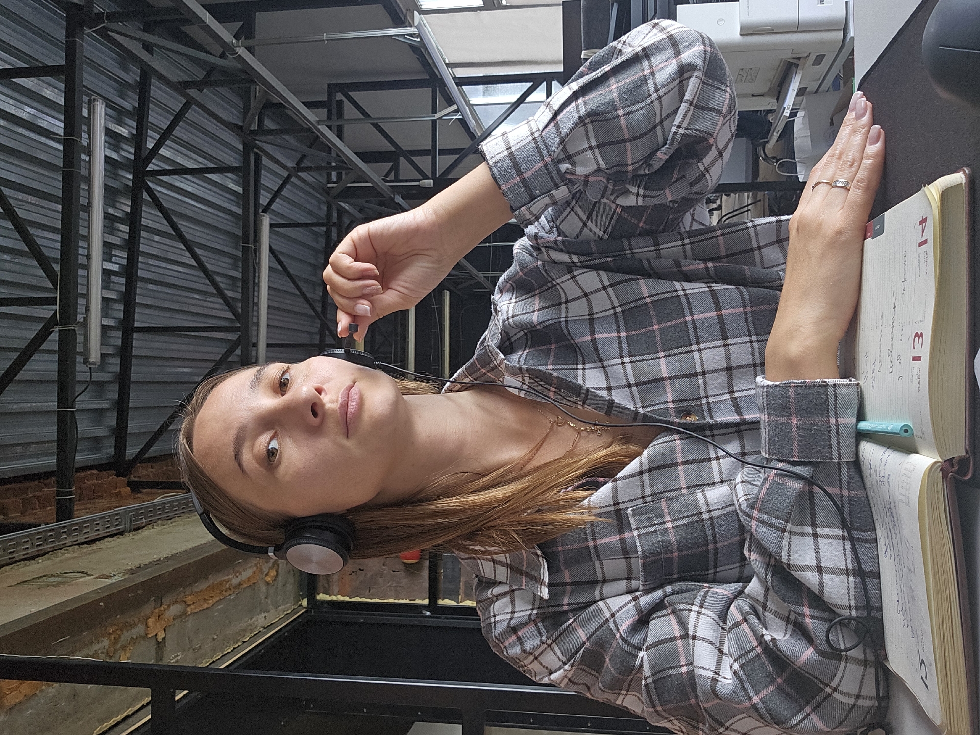 A young woman with light brown hair is sitting at a desk, wearing headphones and a plaid shirt. She is holding a small black object to her ear with her right hand. In front of her is an open notebook with a blue pen. The background shows an industrial-style ceiling with metal beams and fluorescent lights, suggesting an office or workshop environment. The image does not contain any specific indicators to place it in the city of Chaikovsky, Russia.