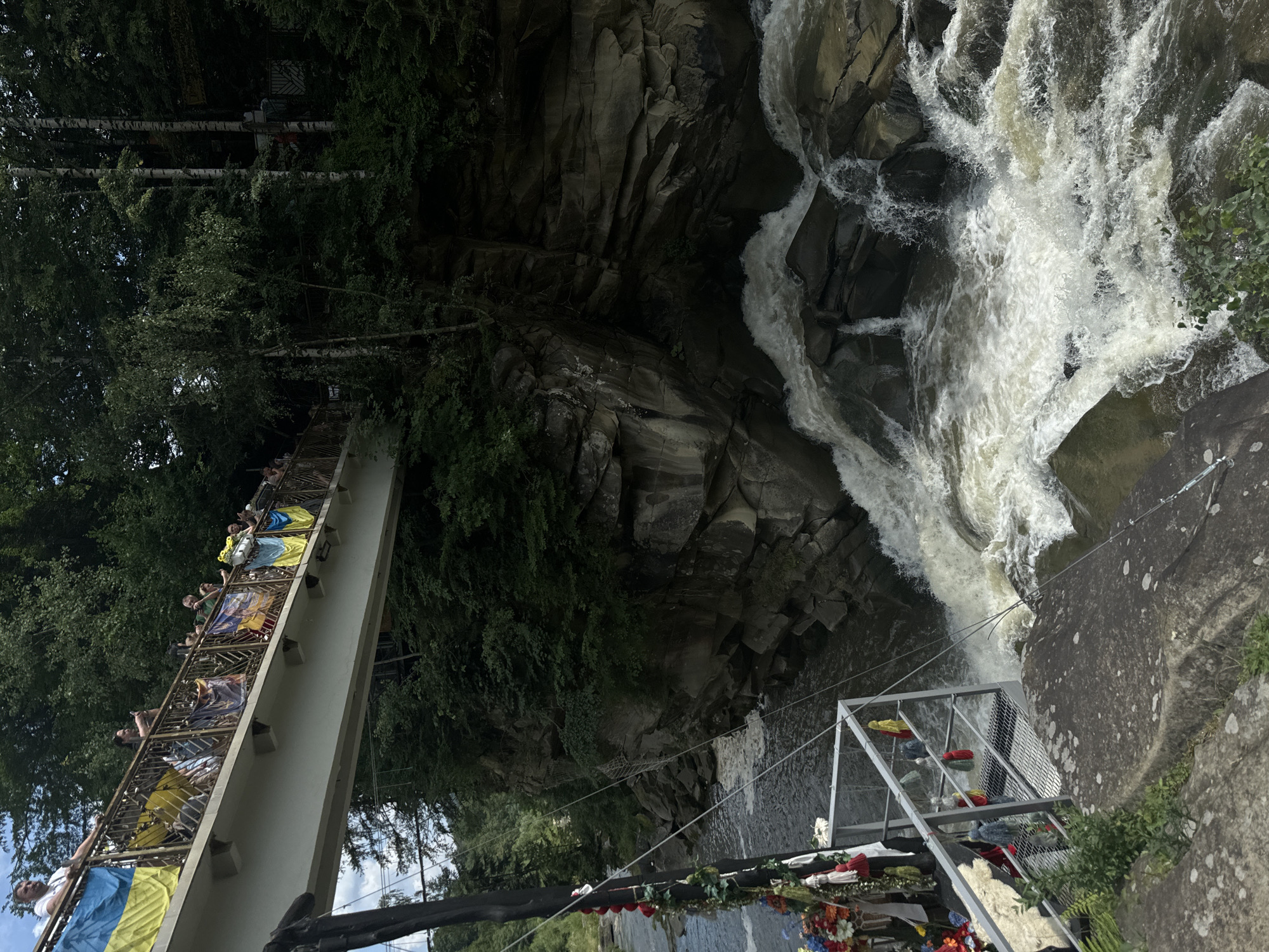 The image shows a view of the Prut River waterfall in Yaremche, Ukraine. A pedestrian bridge spans the river, adorned with numerous Ukrainian flags and banners. Several people are visible on the bridge, some looking at the waterfall and others taking photos. The waterfall cascades over large, dark rocks, surrounded by lush green trees and foliage. The water is turbulent and foamy, indicating a strong current. The foreground includes a metal railing and a rocky outcrop. The overall scene depicts a natural landscape with a man-made structure and a gathering of people.