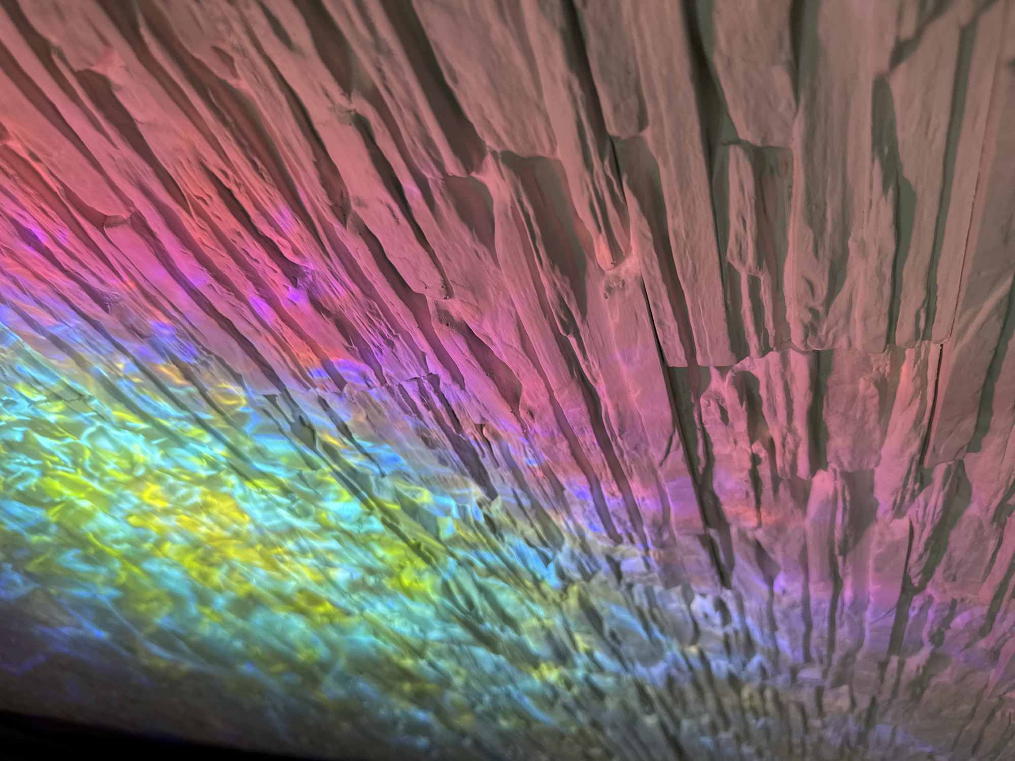 This is a close-up abstract image, with a layered effect. The background appears to be a textured surface with vertical ridges, possibly made of stone or a similar material. The top of the image is dominated by colors ranging from light gray to light pink and purple. The bottom section is more vibrant, with hues of yellow, green, and blue, giving the impression of light reflecting or refracting off a watery surface. The photograph appears to be taken from an unusual angle, with an upward-facing perspective. The specific location in the city of Unknown, Unknown, is not identifiable from the image.