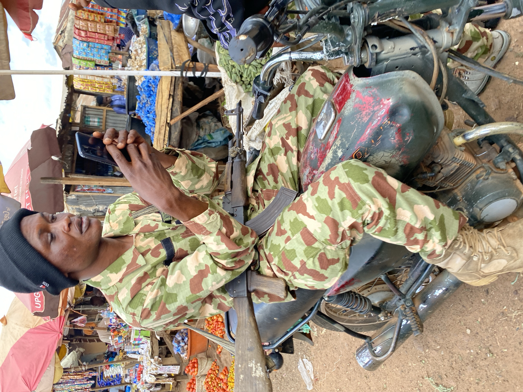In the city of Bukuru, Nigeria, a man in camouflage fatigues and a black beanie sits on a motorcycle. He is looking at his phone, which he holds in his hands. A rifle is resting on the motorcycle in front of him. The background shows a bustling market with stalls selling various goods, including produce and packaged items. The ground is dirt.