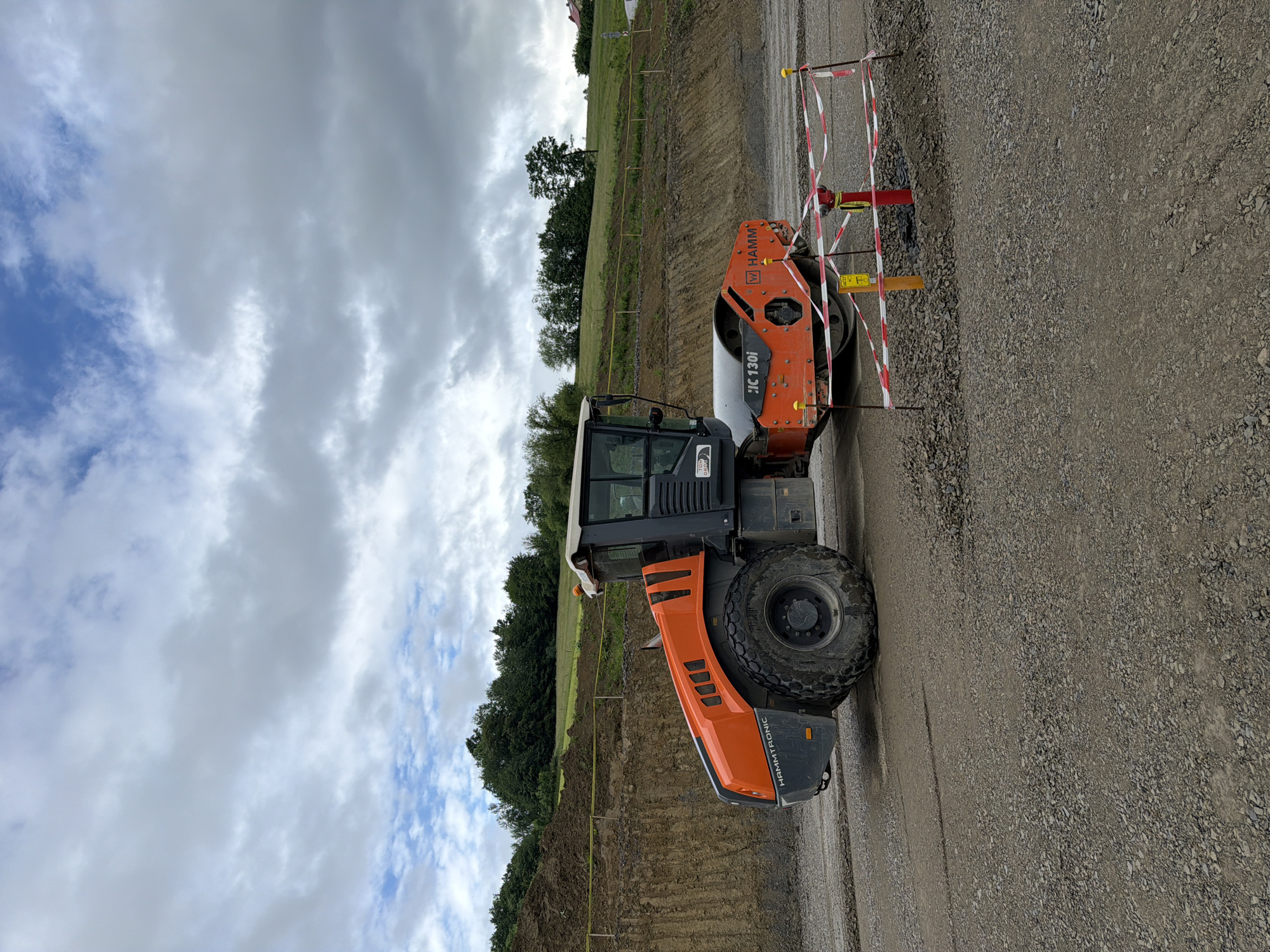This is a medium-shot, eye-level, outdoor photograph of an orange and gray Hamm roller at a construction site, possibly in Janowice, Poland. The roller is positioned on a gravel surface, with a steep, excavated dirt embankment behind it. A yellow caution tape runs horizontally along the embankment. The sky is overcast with scattered clouds. In the foreground, a red and white striped temporary construction barrier with yellow posts surrounds a red fire hydrant and a yellow marker. The roller's tires are large and heavy-duty, and its "HAMM" and "JC 130i" logos are visible. The scene is devoid of people, and the overall impression is one of ongoing construction work.