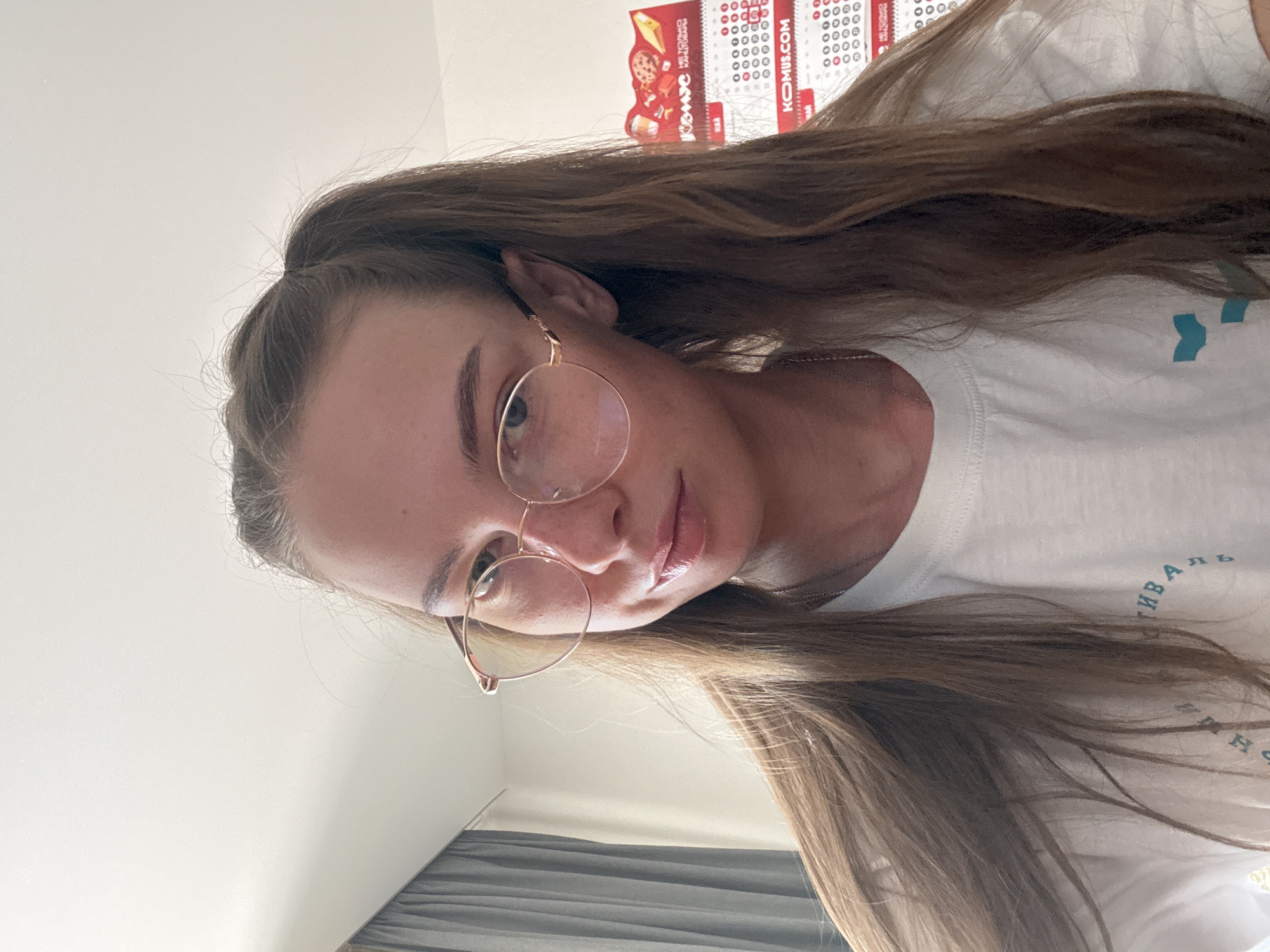 The image is a close-up, selfie-style shot of a young woman with long, brown hair tied in two pigtails. She is wearing round, rose-gold colored glasses and a white t-shirt with a light blue graphic and text. She has light skin and appears to be indoors, with a gray curtain and a calendar visible in the background. The calendar has Russian text and appears to be for the year 2023, with "KOMUS.COM" prominently displayed. The overall lighting suggests an indoor environment, possibly a home or office. There is no visual information within the image that can definitively place the location in the city of Perm, Russia.