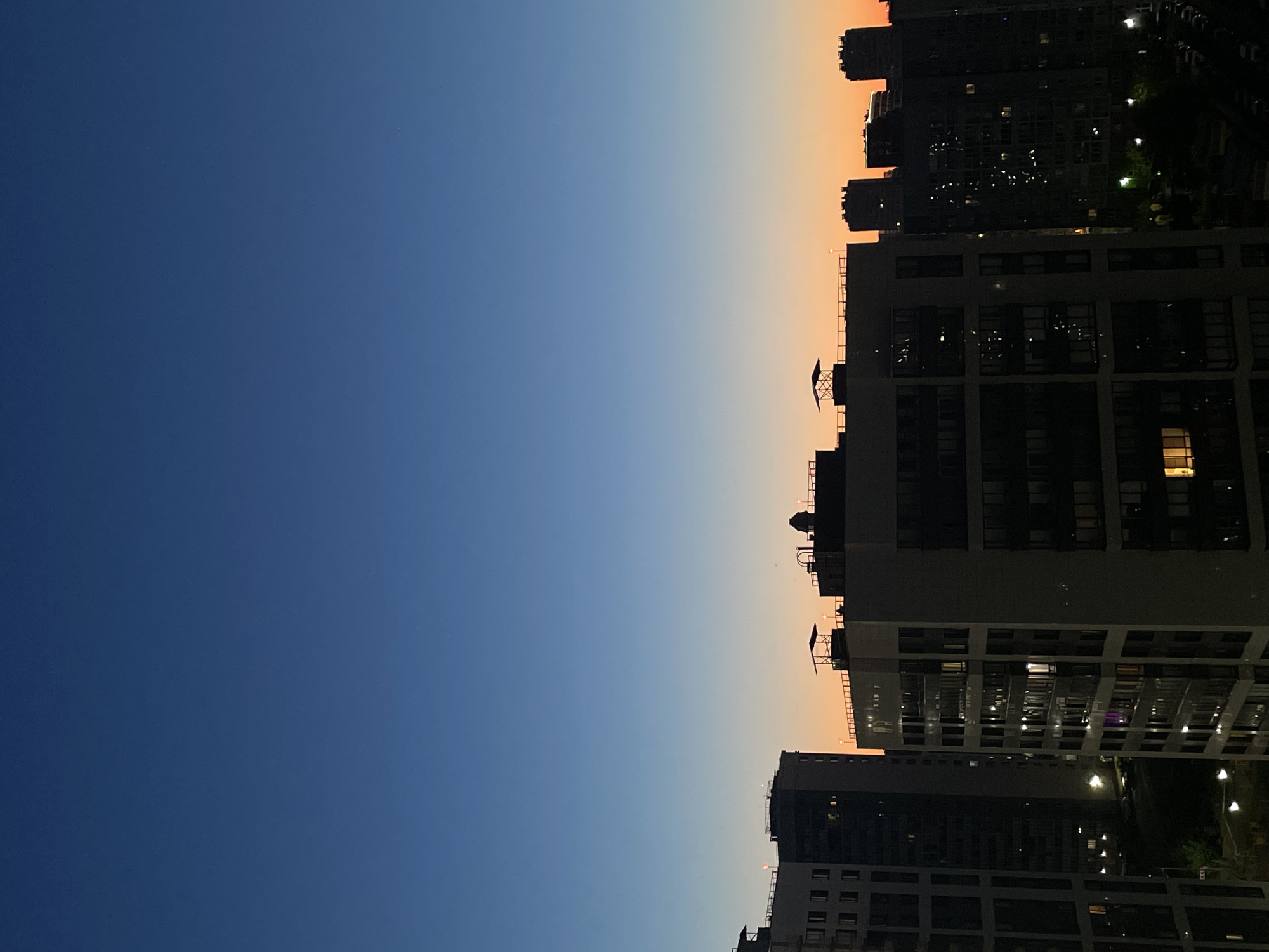 The image depicts a cityscape at dusk in Sinyavino, Russia. Tall apartment buildings dominate the foreground and midground, silhouetted against a gradient sky that transitions from deep blue at the top to a warm orange and yellow near the horizon. Windows of the buildings are illuminated, indicating residents are inside their homes. The buildings themselves appear to be modern residential structures with numerous floors and balconies. In the distance, more buildings of varying heights are visible, also dark against the twilight sky. The overall atmosphere is quiet and serene, capturing a moment as day turns into night. There are no visible people or explicit actions taking place. The environment is urban, characterized by the density of residential construction.