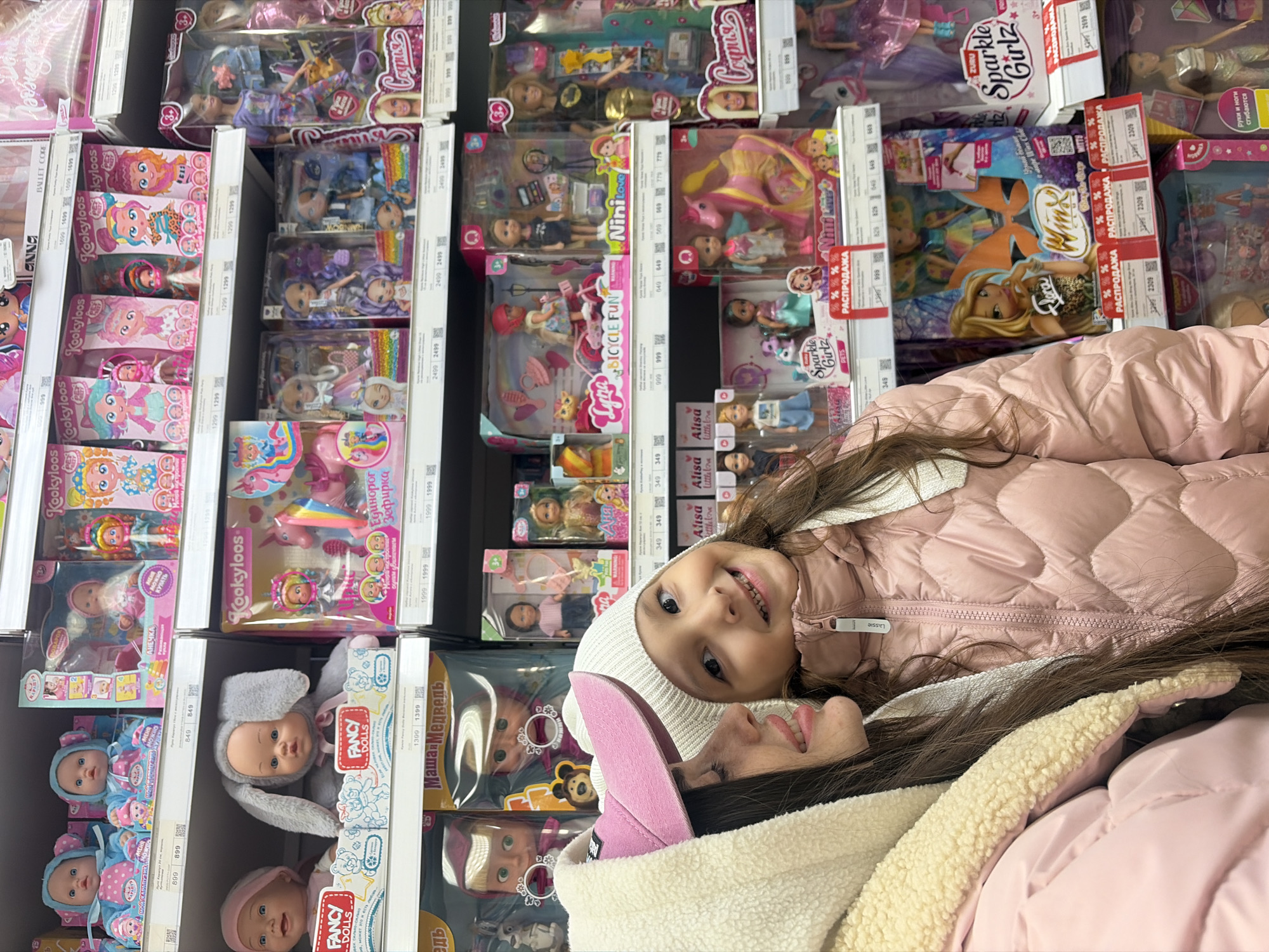 The image displays an interior view of a retail store aisle, specifically a section dedicated to children's dolls and toys. The scene features two individuals in the foreground, partially obscuring the merchandise. A young girl, centrally positioned, faces forward and smiles directly at the camera. She wears a white knit beanie and a pink, quilted puffer jacket with a zippered front. To her left, an adult female is partially visible in profile, wearing a cream-colored, sherpa-lined hooded jacket over a pink garment and a pink visor cap. She has long, dark hair.
Behind the individuals, white multi-tiered shelving units are packed with various boxed dolls and toy sets. Notable toy brands and types include "Fancy Dolls," "Kookyloons," "Masha i Medved" (Masha and the Bear), "Lyra Bicycle Fun," "Nini Love," "Sparkle Girlz," and "Winx Club" dolls. Price tags with figures in Russian Rubles (e.g., 899, 1299, 2499) and Cyrillic text such as "РАСПРОДАЖА" (SALE) are visible on the shelf edges. The overall environment is brightly lit, consistent with a commercial retail space. While the presence of Cyrillic script and Russian product brands confirms a retail establishment in Russia, the precise location within the city of Saint Petersburg is not ascertainable from the visual information provided.