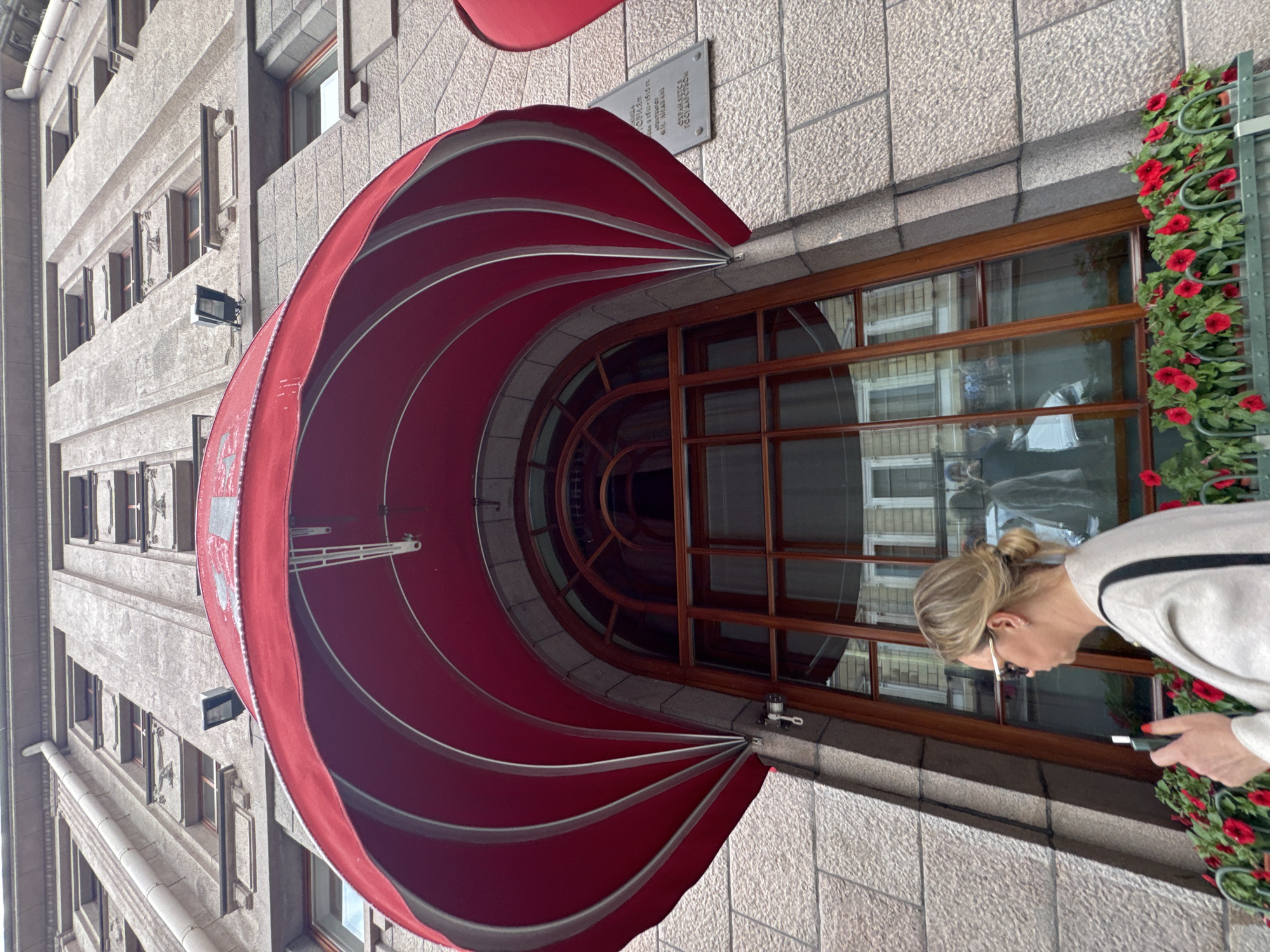 The image shows a low-angle view of the facade of the Singer House (Dom Knigi) in Saint Petersburg, Russia, a landmark building located at Nevsky Prospekt, 28. The building features a stone facade with multiple windows and a prominent red awning over a large arched window. A woman with blonde hair pulled into a bun and wearing sunglasses is visible in the foreground, holding a white electronic device. She is standing in front of the arched window, which reflects the surrounding buildings and street. Red flowers are planted in planters along the base of the window. A historical plaque is visible on the building's facade to the right of the window. The architecture suggests an early 20th-century style, characteristic of buildings on Nevsky Prospekt.