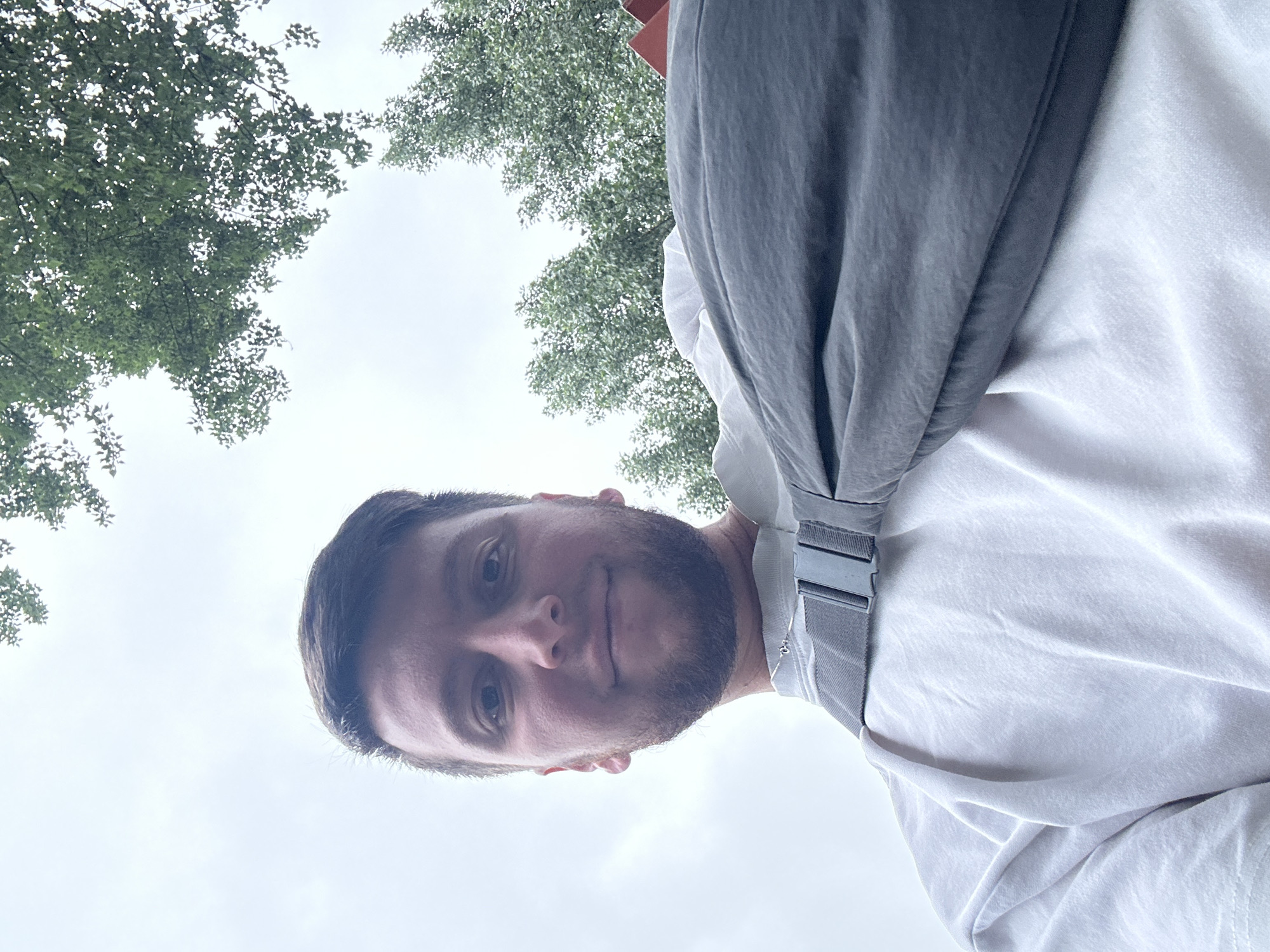 A low-angle, close-up, outdoor, waist-up selfie shows a young man with short brown hair and a beard, smiling at the camera. He is wearing a white t-shirt and a dark gray sling bag across his chest. The bag has a gray strap and a black buckle. The background consists of a cloudy sky and green trees. The image does not contain any information to suggest the location is Moscow, Russia.