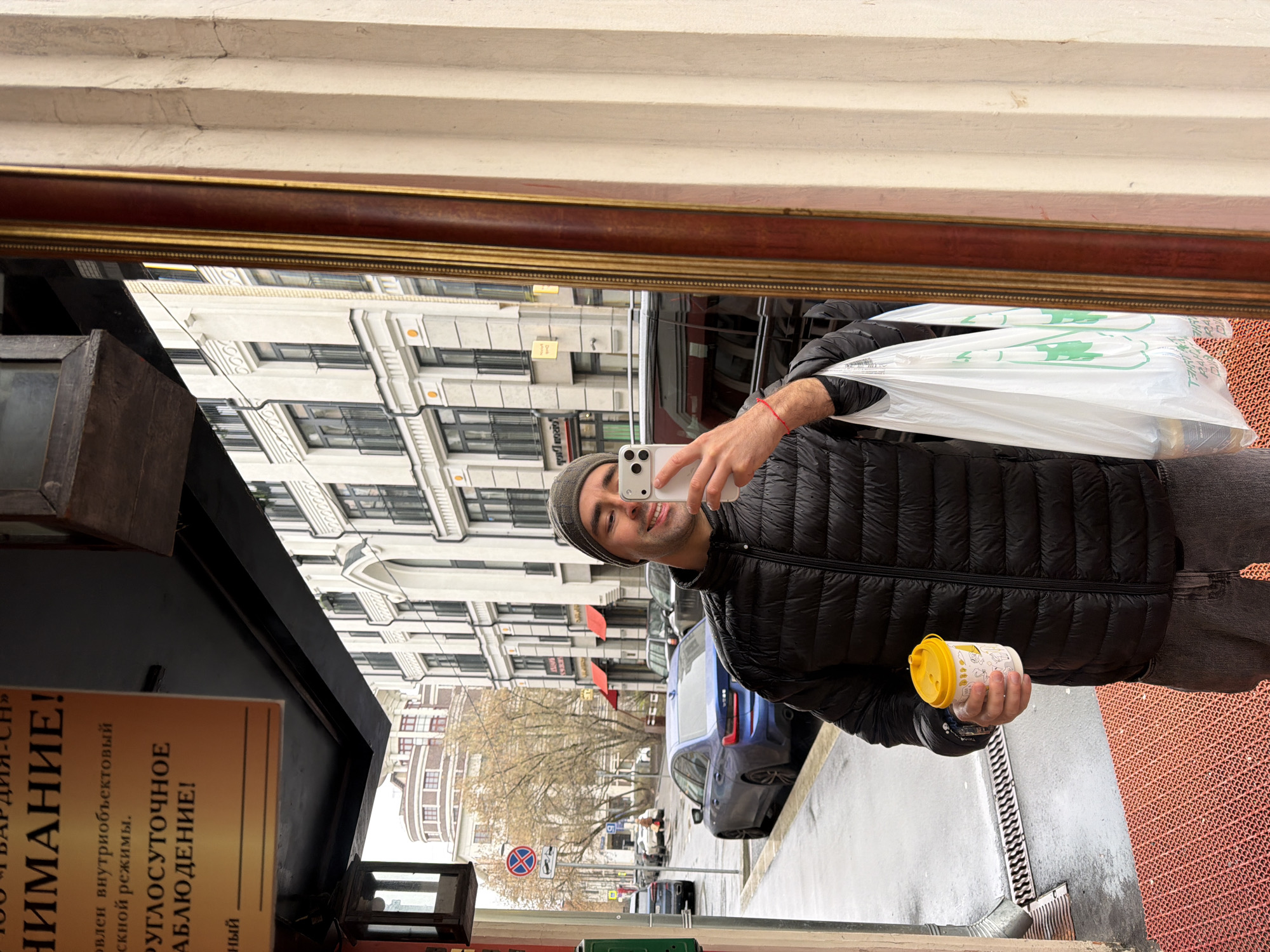 Here is a concise description of the image:
The image is a selfie taken in a mirror's reflection on a street in Moscow, Russia. A man wearing a dark jacket and a beanie holds a smartphone in one hand and a small yellow container in the other. He also carries a white plastic bag.
The reflection shows a street scene with multi-story buildings, parked cars (including a blue BMW), and street signs. The buildings appear to be of European architectural style.
The mirror is set within a frame and reflects a storefront sign with a Russian inscription. The sign includes the word "Внимание!" (Attention!).