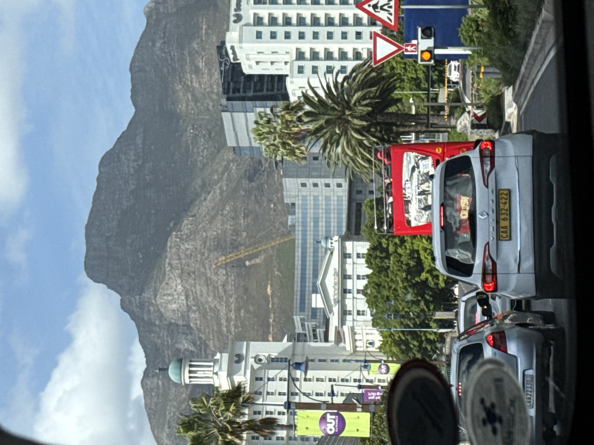 The image shows a view from a vehicle in Cape Town, South Africa. The prominent feature is Table Mountain in the background, with cloudy skies above. In the foreground, there are several buildings, including one with a dome. A traffic light is illuminated in yellow. The road is filled with cars in traffic. The vehicle directly in front has license plate CAA 632-422. A red bus is next to it. A traffic sign with a person and a triangle is visible. There are palm trees. A sign for 'OUT' is apparent near a building. A crane is working on a building site.