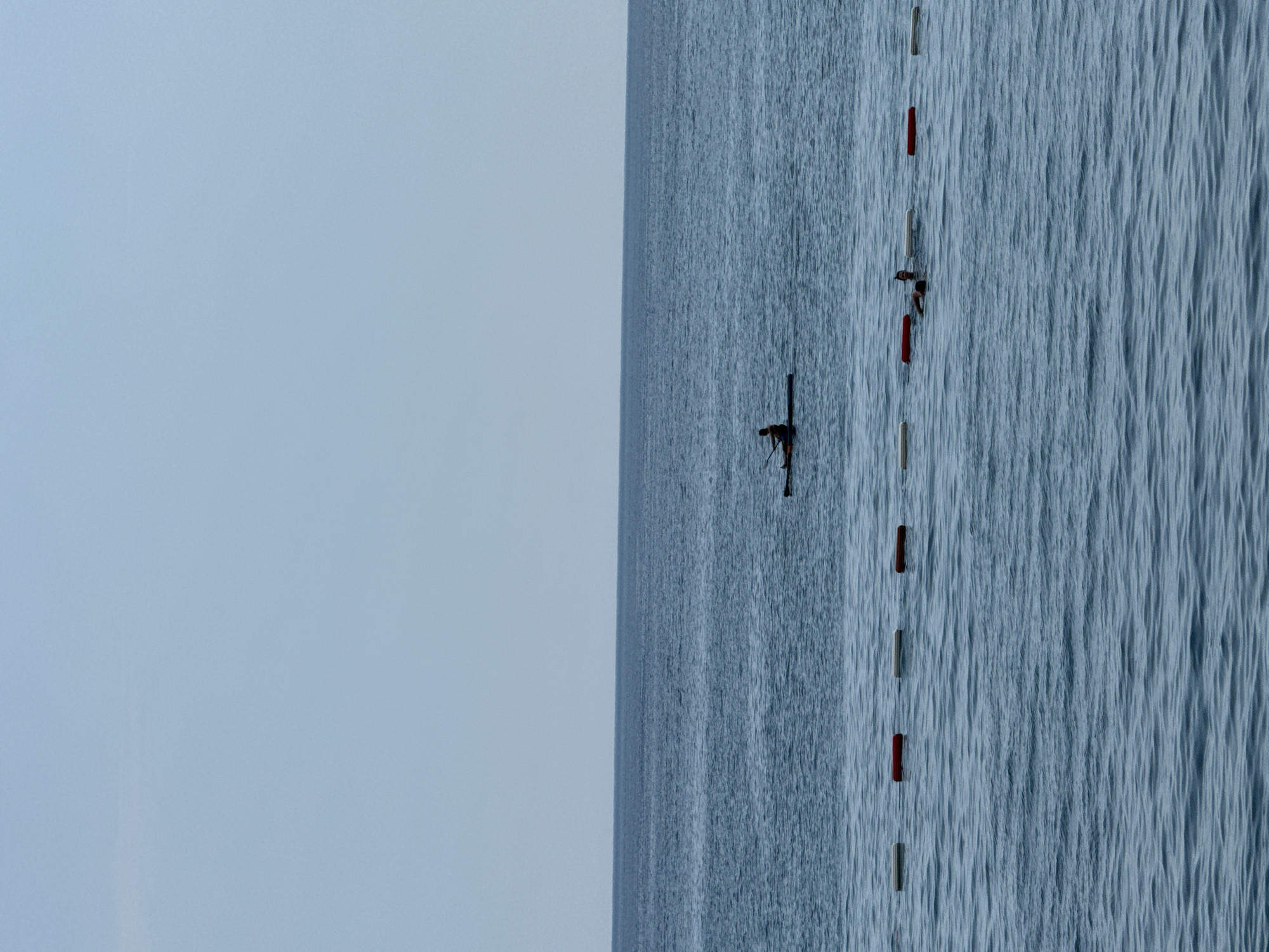 The image shows a wide expanse of calm, blue ocean under a clear, pale blue sky. The horizon line where the sea meets the sky is sharp and distinct. In the mid-ground, a person is silhouetted on a paddleboard, paddling through the water. Further towards the foreground, a series of buoys, alternating between white and red, mark a boundary in the water. Behind these buoys, two individuals are partially submerged in the sea, appearing to be swimming or floating. The overall scene depicts a serene day at the coast, likely in the city of Rafailovići, Montenegro, with recreational activities taking place on the water.