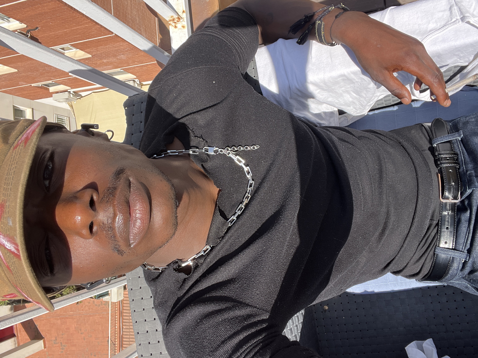 This image captures a person in a bright, sunny outdoor setting, likely a rooftop terrace or balcony in Barcelona, Spain. The subject is a person, appearing male, with dark skin, looking directly towards the camera. They are dressed in a black long-sleeved shirt, blue jeans, and a black belt. A silver chain necklace with rectangular links is prominent around their neck. On their right wrist, multiple bracelets, some featuring tassels, are visible. A tan or khaki baseball cap with a subtle red detail covers their head.
The person is in a relaxed pose, with their right arm resting casually. Strong sunlight illuminates the scene, casting distinct shadows, indicating a clear, bright day, likely mid-morning or afternoon. The background features architectural elements characteristic of a Mediterranean city, including red-tiled rooftops, white horizontal beams of a pergola-like structure, and a yellow building facade with windows. The bright conditions and urban architectural details suggest a warm, pleasant atmosphere. No visible text is present in the image.