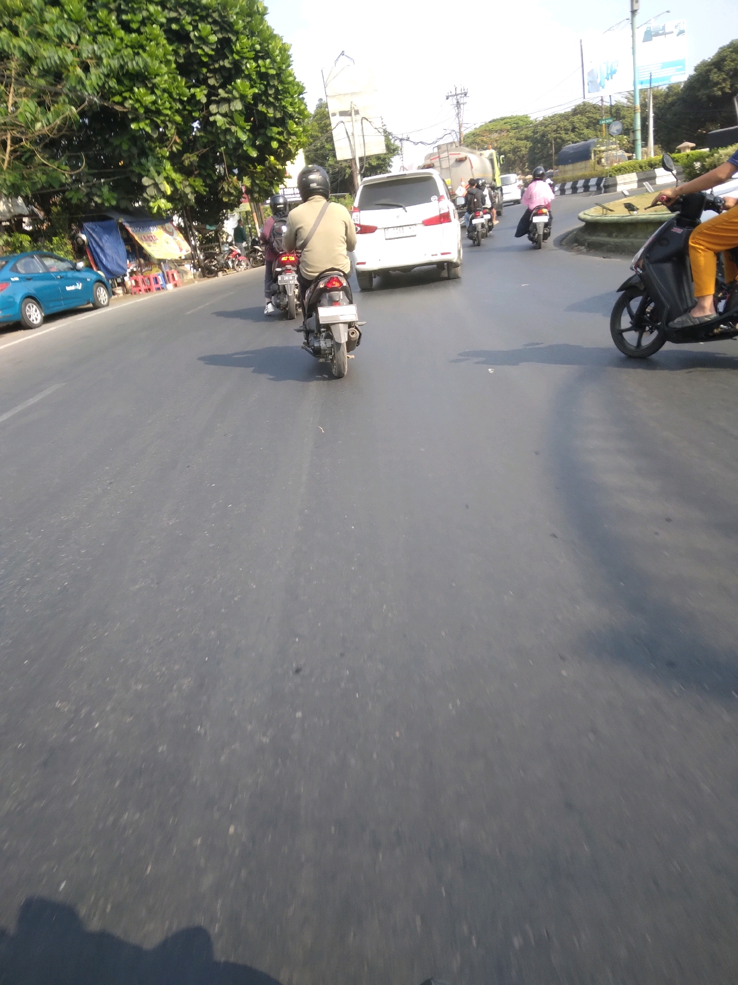 The image depicts a street scene in Sokaraja, Indonesia, on a sunny day with clear skies. The road is paved and appears to be a multi-lane street with moderate traffic. In the foreground, a person on a dark-colored scooter is moving forward, casting a shadow on the road. The person is wearing a light-colored long-sleeved shirt and dark pants, with a helmet on their head.
Further down the road, several other vehicles are visible. To the left, a blue car is parked on the side of the road. Behind the blue car, there are some stalls or small shops with blue tarps and red and yellow accents. Several motorcycles are parked in front of these establishments.
In the middle distance, a white car is driving away from the camera. Behind it, a person in a pink shirt is riding a motorcycle. Other motorcycles and cars are scattered throughout the scene, indicating ongoing traffic flow.
The environment includes lush green trees lining the side of the road, with some trees obscuring parts of the background. Electrical poles and wires are also present, typical of urban or semi-urban environments. In the far distance, there are more trees and some buildings, suggesting a developed area. The overall impression is a busy street in Indonesia during daylight hours.