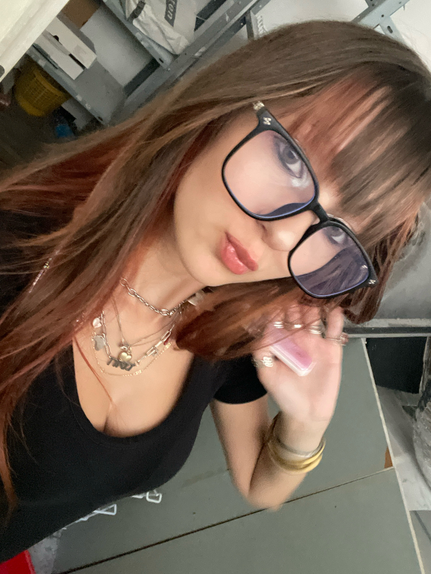 A woman with long, brown hair streaked with pink is wearing a black V-neck top and large, black-rimmed glasses. She has on multiple necklaces, including one with a heart pendant, and several rings on her fingers. She is holding a small, pink rectangular object in her right hand. The background appears to be a cluttered indoor space, possibly a storage area or workshop, with shelves and various objects visible. The image does not provide any specific information that would allow for identification of a location in the city of Raanana, Israel.