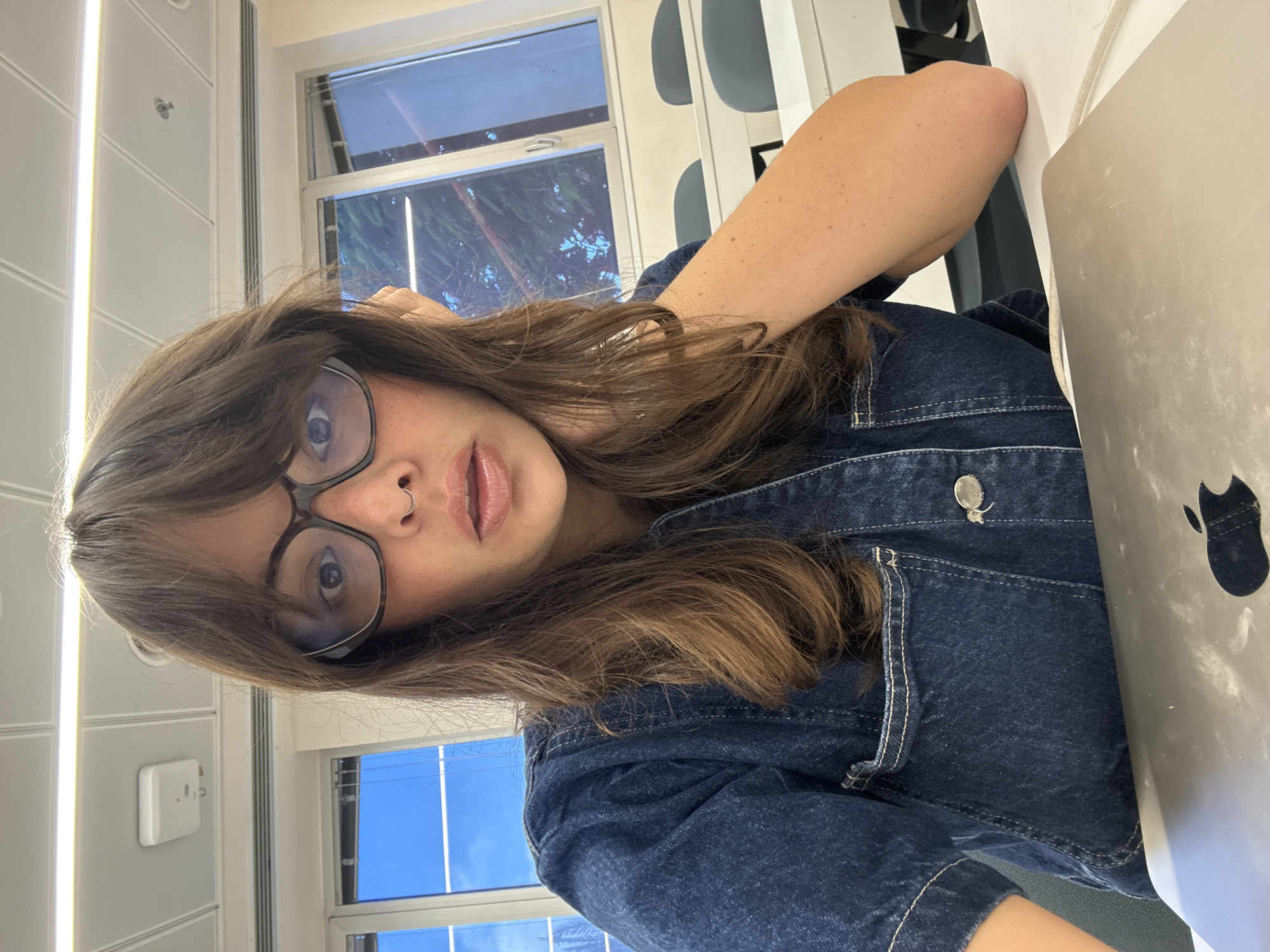 A young woman with long brown hair and bangs, wearing glasses and a denim shirt, is seated at a desk in front of a laptop. Her right arm is resting on the desk, and her hand is supporting her head. She is looking directly at the camera with a neutral expression. The background appears to be a classroom or lecture hall, with rows of empty desks and chairs, large windows, and fluorescent lighting. The image was taken indoors.