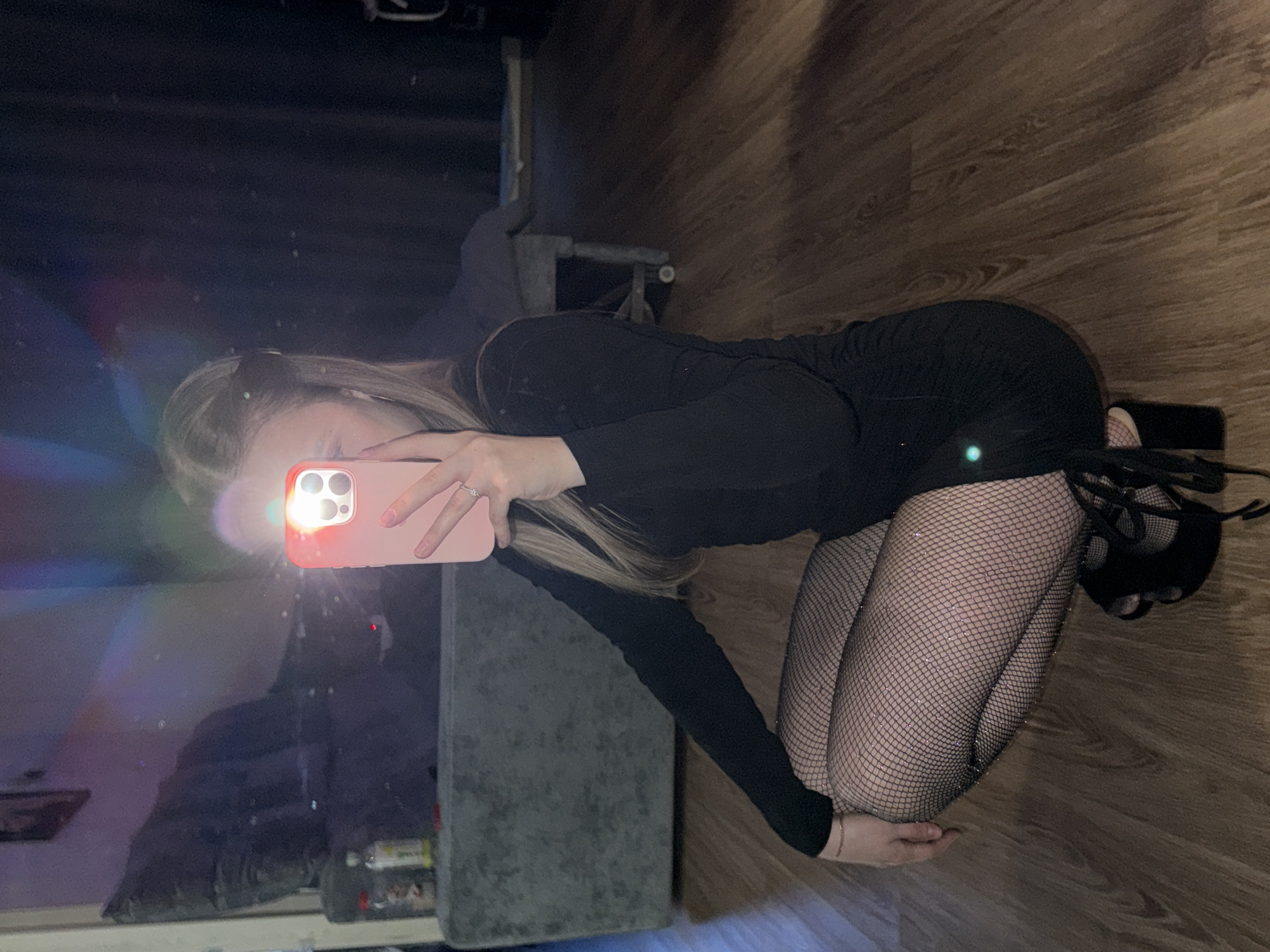 An indoor mirror selfie captures a young woman kneeling on a wooden floor in Chaikovsky, Russia. The image is oriented with the floor on the right, and the woman's body spans the frame vertically.
The subject has long, blonde hair and is wearing a black long-sleeved dress or top. She is also wearing shimmering fishnet stockings and black high-heeled shoes with laces. She is positioned on her knees, leaning forward, and holding a smartphone with a pink/peach case in front of her face. The phone's flash is active, creating bright lens flares and reflections, particularly on the left side of the image. A silver ring is visible on her left hand.
The flooring is made of wood, displaying a visible grain pattern. The background is dark and indistinct, showing reflections consistent with a mirror selfie. A dark, possibly upholstered piece of furniture or an object with a textured surface is partially visible on the far left. The lighting is dim, suggesting the photo was taken at night or in a poorly lit room, with the phone's flash providing the primary illumination. No text is visible in the image.