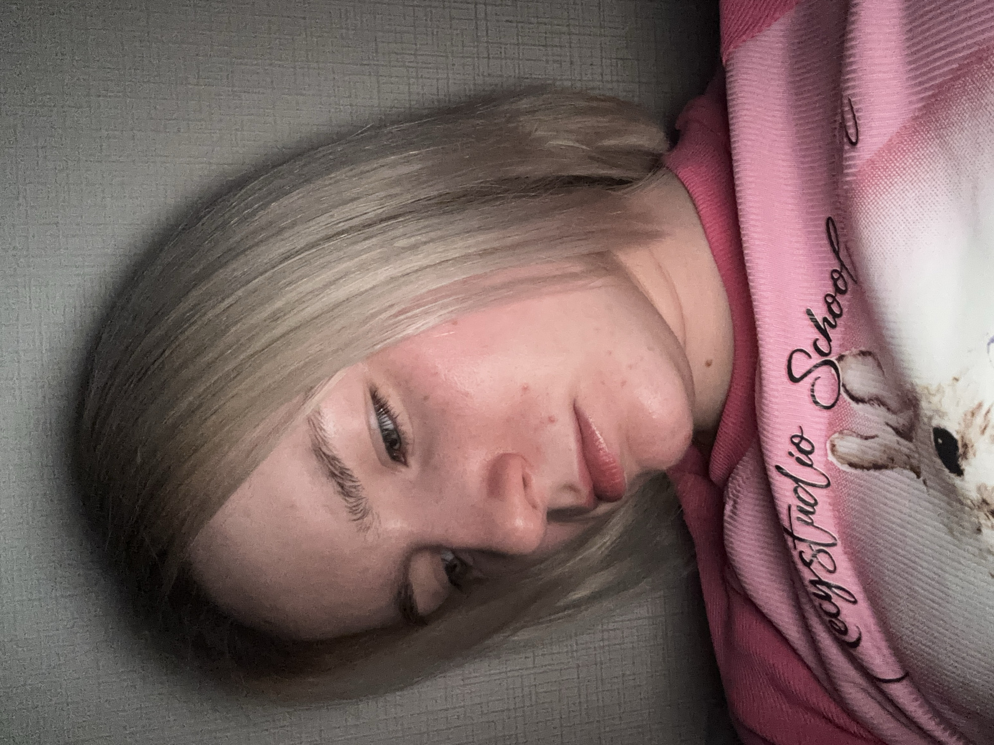 A close-up, tilted portrait shows a young person, likely a woman or girl, with blonde or light brown hair, resting indoors in Ligolambi, Russia. Her head is tilted to the right, with her face looking downwards and to the left. Her eyes are open, and her expression appears neutral or slightly pensive. Visible on her skin are some small blemishes and areas of redness on her cheek and forehead.
She is wearing a pink garment, possibly a hoodie or blanket, made of a soft, ribbed fabric. The garment features visible black text that reads "ecystudio School," though the initial part "ecy" might be "Lecy" or "Secy" due to partial obstruction. A faint, light-colored design, possibly depicting a cartoon animal like a bunny or cat, is also visible on the garment's right side. The background is a grey, textured wall or surface. The lighting is soft and ambient, characteristic of an indoor setting.
