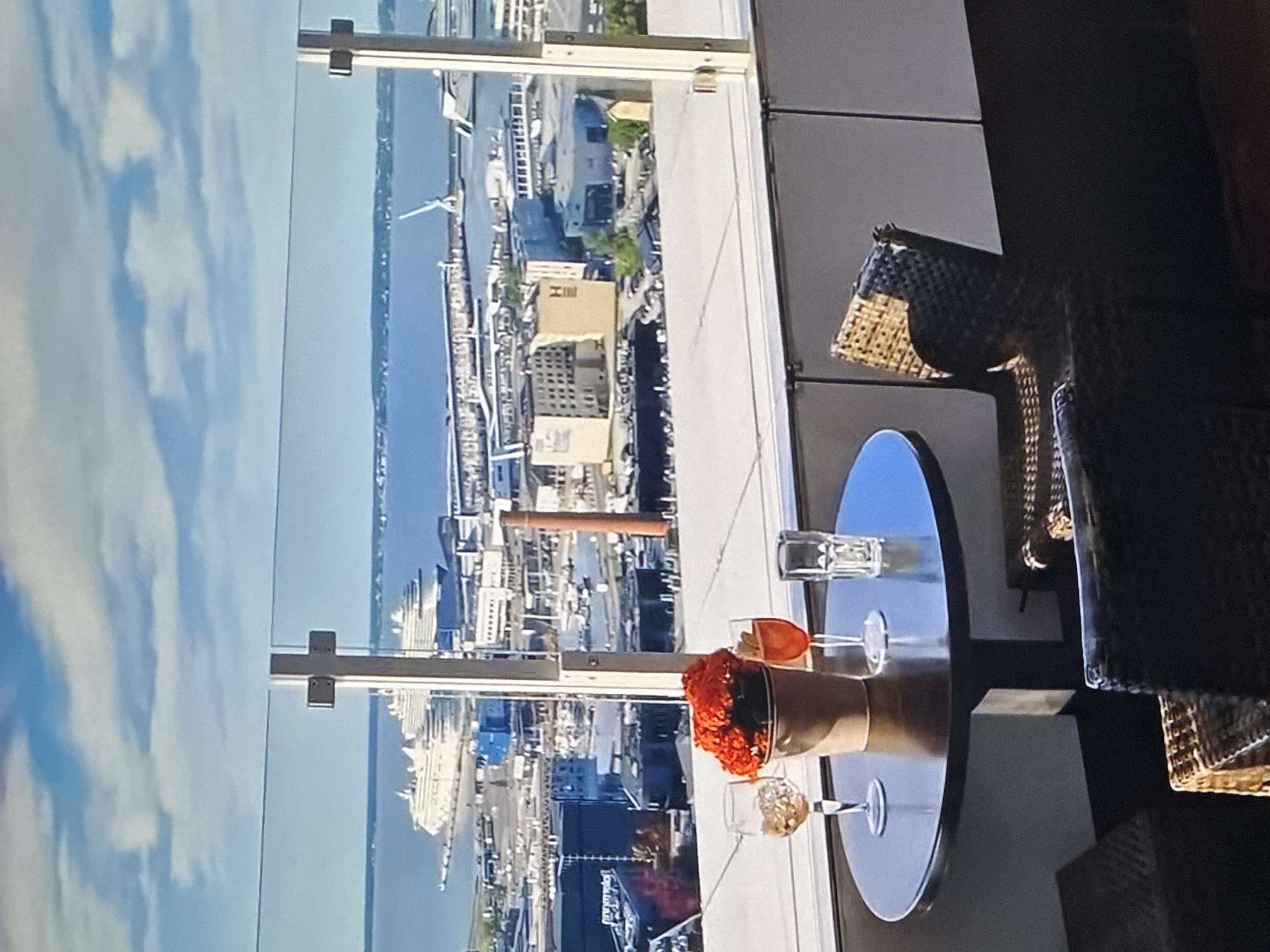 This image captures a view from a high-rise balcony in Tallinn, Estonia, overlooking the city's port. In the foreground, a round dark table holds a bouquet of bright orange flowers in a light brown pot, two stemmed glasses with beverages, and a clear drinking glass. A wicker chair is partially visible to the right of the table. The background features a wide expanse of the Baltic Sea, with several large cruise ships docked in the harbor. Beyond the ships, the city skyline is dotted with various buildings, industrial structures, and cranes, indicative of a busy port area. The sky above is a mix of blue and white clouds, suggesting daytime.
