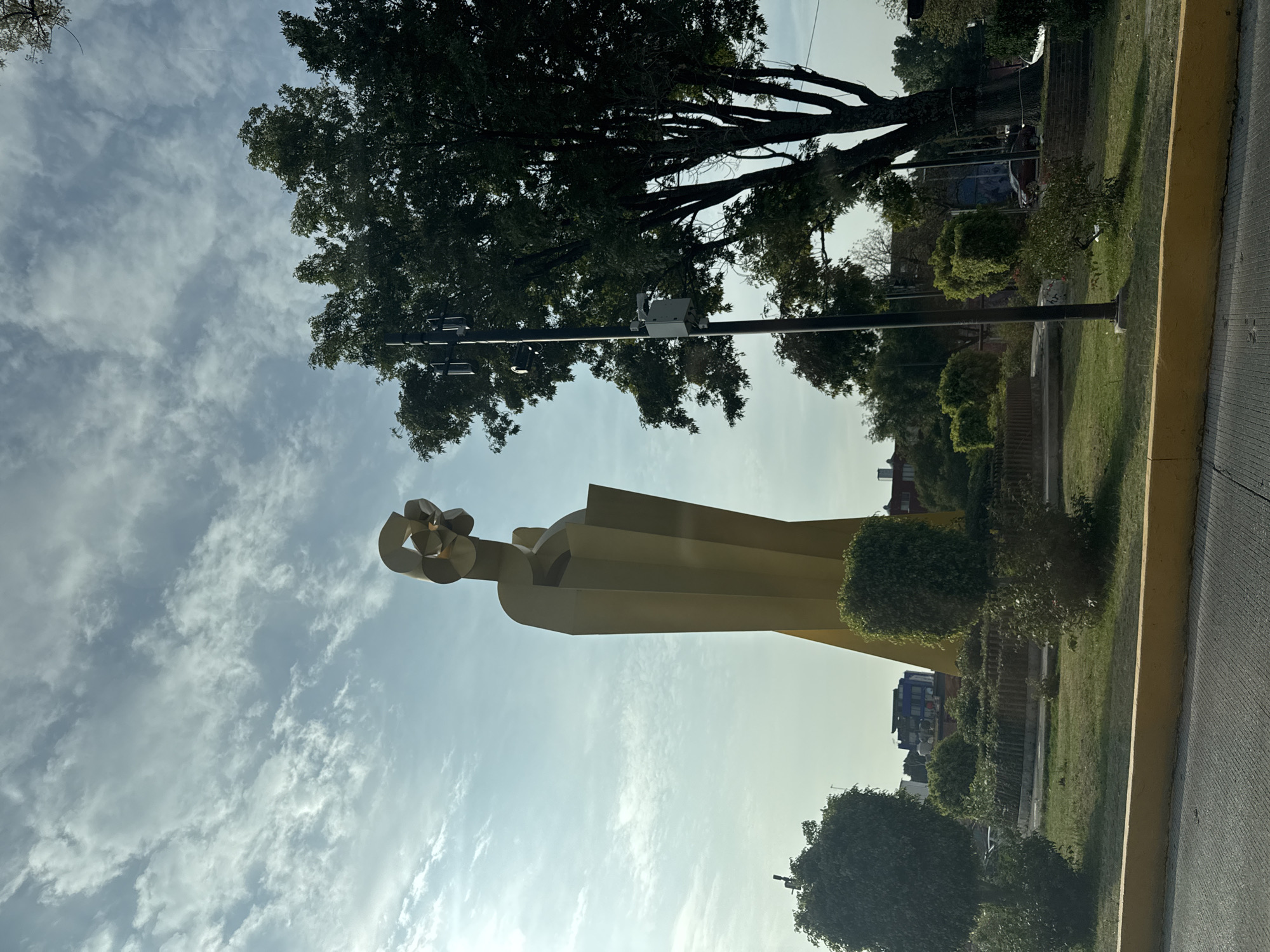 This image, taken in Puebla, Mexico, showcases a large, striking yellow sculpture set against a bright, partly cloudy sky. The sculpture is abstract, featuring a stylized figure with a raised arm that holds a multifaceted, circular object composed of interconnected geometric shapes. Its form is blocky and architectural, pointing upwards towards the sky.
The sculpture is situated within a landscaped green area, likely a park or a wide median strip. This area is covered with green grass and features various trees and manicured bushes. In the background, darker green trees with prominent branches line the horizon, suggesting a mature and established natural environment.
The sky above is a clear, light blue with scattered white clouds, indicating a daytime scene with fair weather conditions. To the right, a grey asphalt road or pathway is visible, bordered by a distinct yellow curb, implying the presence of a street or public thoroughfare. A utility pole, equipped with what appears to be a surveillance camera or sensor, stands to the right of the sculpture, further integrating the scene into an urban or suburban context. Faint outlines of distant buildings can be seen beyond the trees. No human activity or discernible text is present in the static scene.