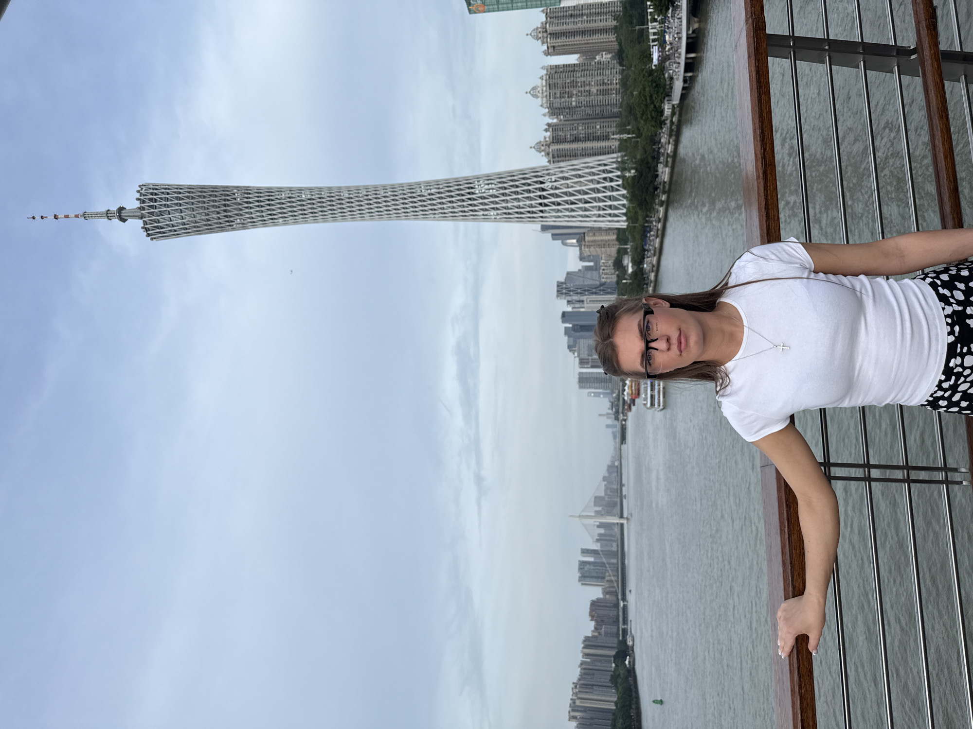A woman stands on a bridge overlooking a river and a city skyline. She is wearing a white t-shirt and a patterned skirt. In the background, the Canton Tower, a landmark in Yuexiu District, China, is visible. The sky is overcast, and there are several boats on the river. The woman is facing the camera with a neutral expression.