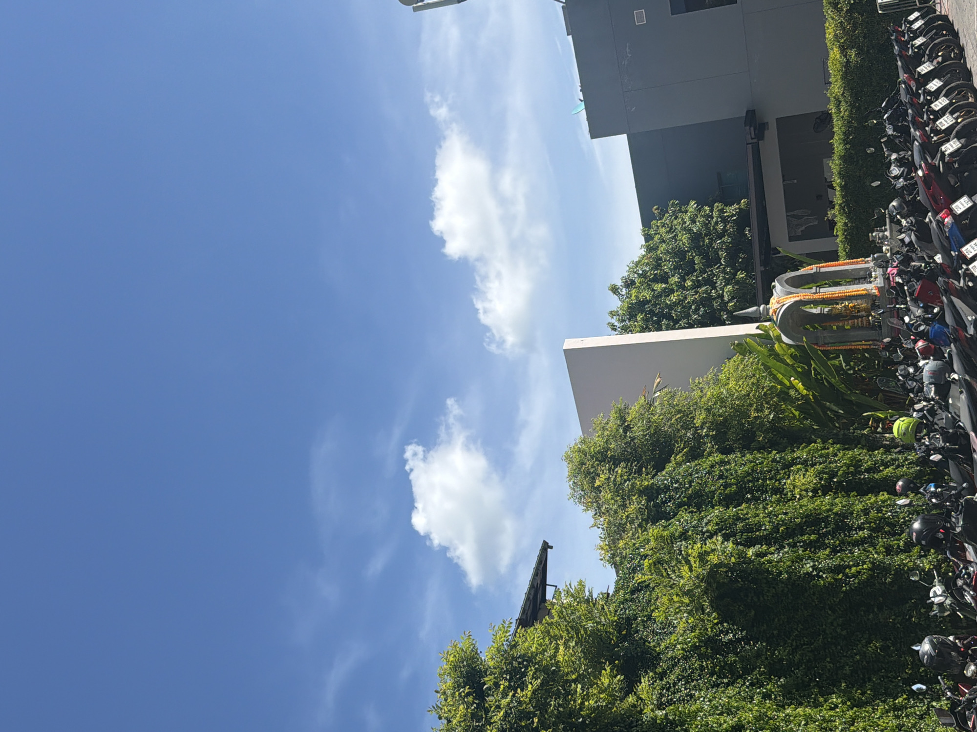A sunny, outdoor scene in Baan Chaweng, Thailand, features a parking lot filled with numerous motorcycles. The foreground is dominated by a dense, green, ivy-covered wall, with the tops of several motorcycles visible below it. To the left, a building with a dark, sloped roof extends partially into the frame, also covered in green foliage. In the center of the image, a small, ornate shrine with arched openings and decorative garlands stands in front of a low, manicured hedge. Behind the shrine, a multi-story building with a gray facade is visible, featuring dark windows and a prominent antenna on its roof. The sky above is a clear, bright blue with scattered white clouds.