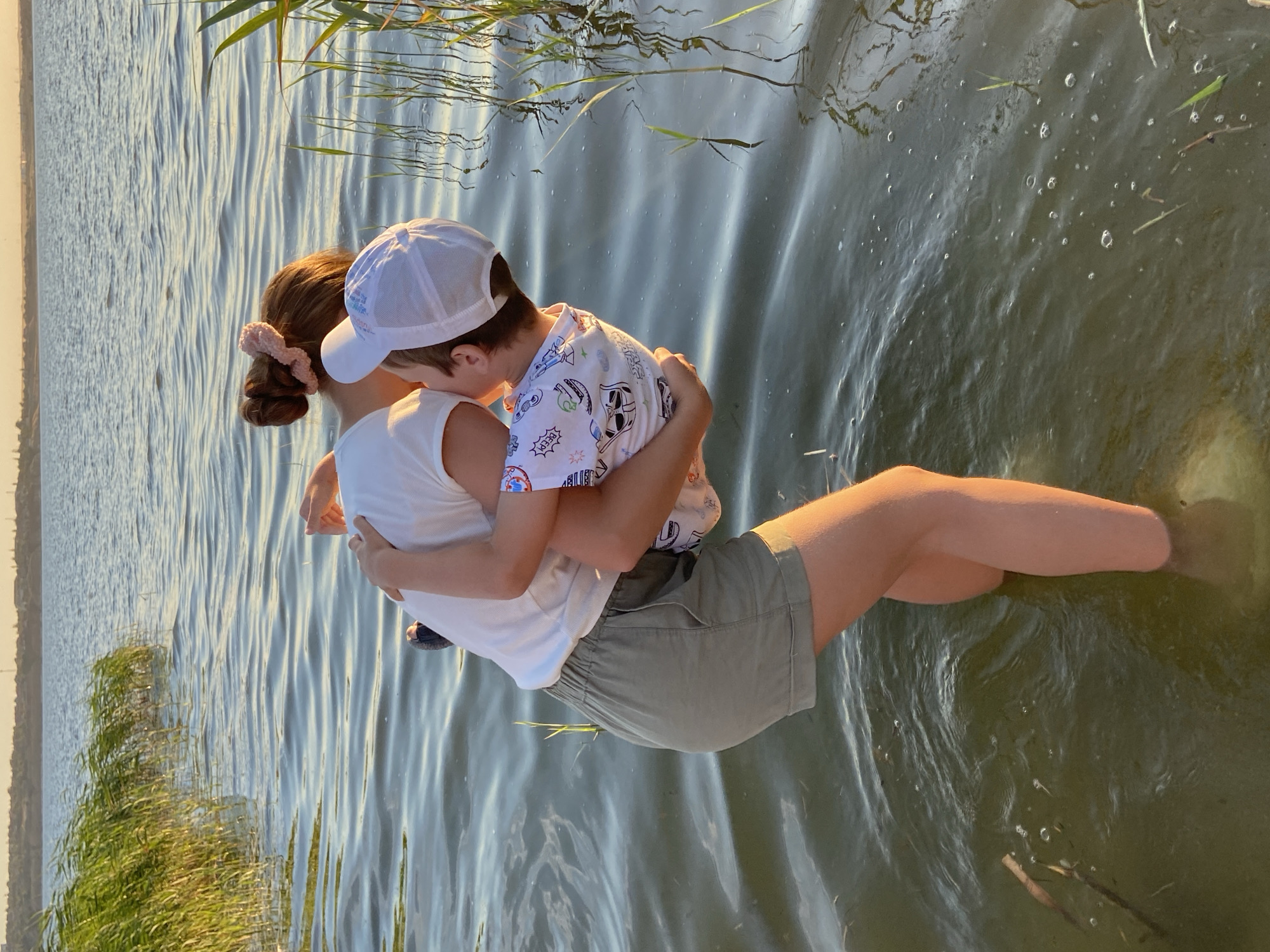 The image shows two people, likely a child being held by an older individual, in the water. The child is wearing a white baseball cap and a white t-shirt with a playful, cartoon-like pattern. The person holding the child is wearing a white t-shirt and olive-green shorts. They are positioned with their backs to the viewer, and the child is facing them, embracing them. Their legs are submerged in the water, and ripples indicate movement.
The scene appears to be a natural body of water, possibly a lake or river, with reeds or tall grass growing along the edge on the left side of the frame. The water is calm, with gentle ripples reflecting the sunlight. The overall lighting suggests it is late afternoon or early evening, with a warm glow on the water and the people. The mood conveyed is one of affection and perhaps playfulness, as the two individuals seem to be enjoying their time together in the water. There is no visible text in the image.