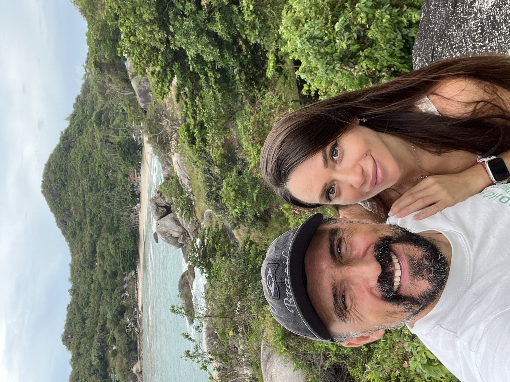 A selfie shows a woman with long brown hair and a man with a salt-and-pepper beard and a dark baseball cap on a rocky coast. They both smile. A sandy beach, ocean water, and a green, densely vegetated hill slope are visible in the background. The scene appears to be in a coastal location near Baan Chaweng Noi, Thailand.