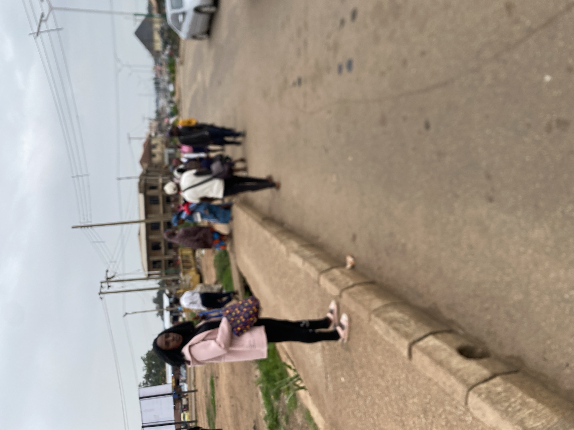 In Bukuru, Nigeria, a young woman in a light pink coat and black leggings stands on a concrete sidewalk, holding a patterned bag. She is positioned on the left side of the frame, facing left with her body angled slightly towards the viewer. Behind her, a group of people are walking along a paved road. The road is lined with buildings and utility poles. The sky is overcast.