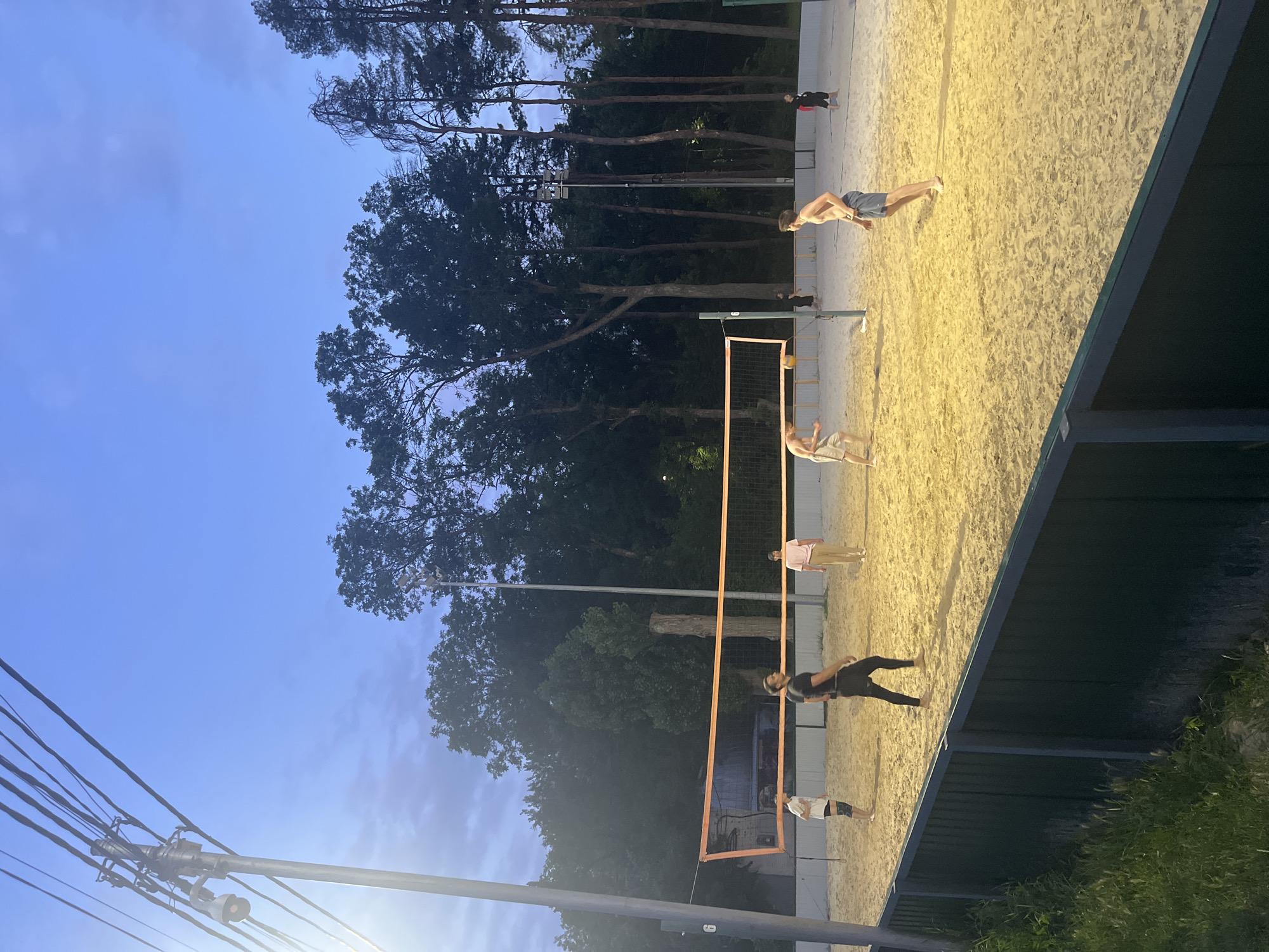This is a photograph of a beach volleyball game being played on a sand court at dusk. The court is illuminated by artificial lights, casting a warm glow on the sand. A net divides the court in half, and a volleyball is in mid-air, suggesting an active game. Several individuals, dressed in casual athletic wear, are participating in the game, with some in motion as if playing. The background shows a line of tall trees, indicating an outdoor setting. The sky above is a deep blue with subtle cloud formations. The scene appears to be located at a public park or recreational area within the city of Krasnodar, Russia, possibly at a sports complex, given the organized setup of the volleyball court.