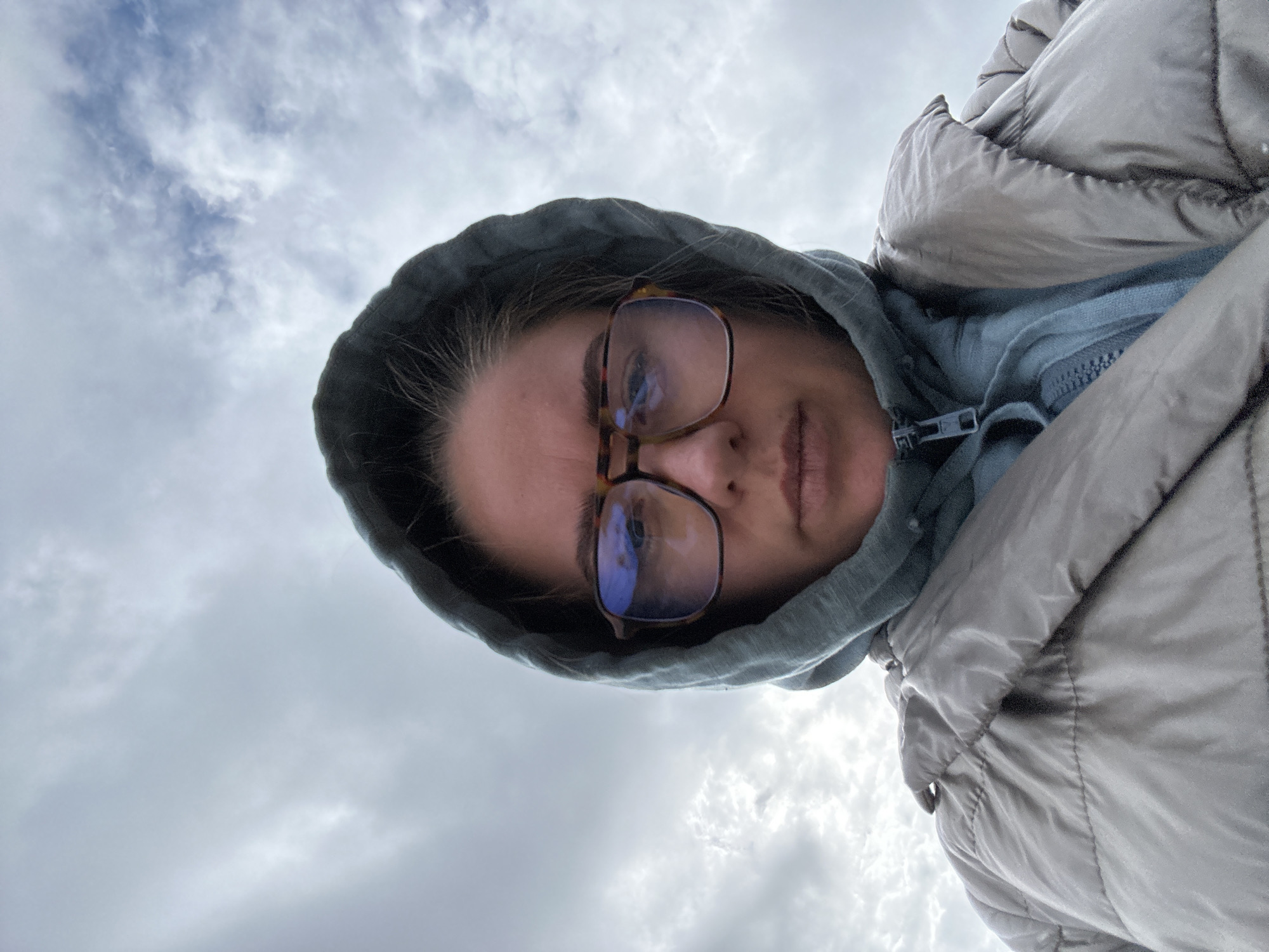A woman wearing glasses, a light blue hooded sweatshirt, and a grey puffer jacket is angled upward towards the sky. The sky is overcast with grey and white clouds. The photograph was taken in a location with cloudy skies.