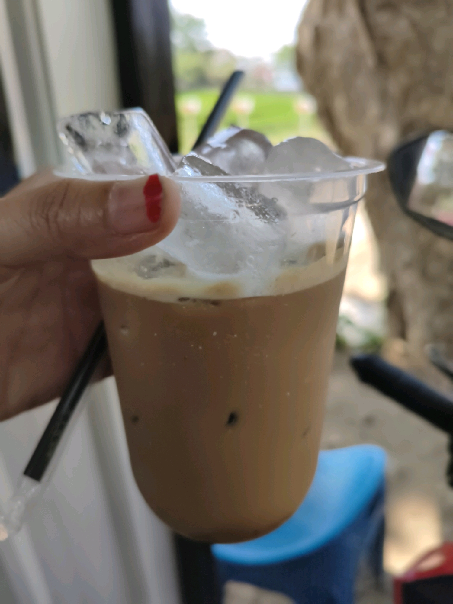 A close-up, shallow depth-of-field shot shows a person's hand holding a clear plastic cup filled with iced coffee. The cup contains a significant amount of ice, with some cubes visible above the coffee's surface. A black straw is inserted into the drink. The person's thumb is visible, with a small red mark, possibly nail polish or a sticker, on the tip of the thumb. The background is blurred, suggesting an outdoor setting in the city of Puhpelem, Indonesia, with hints of greenery and possibly structures.