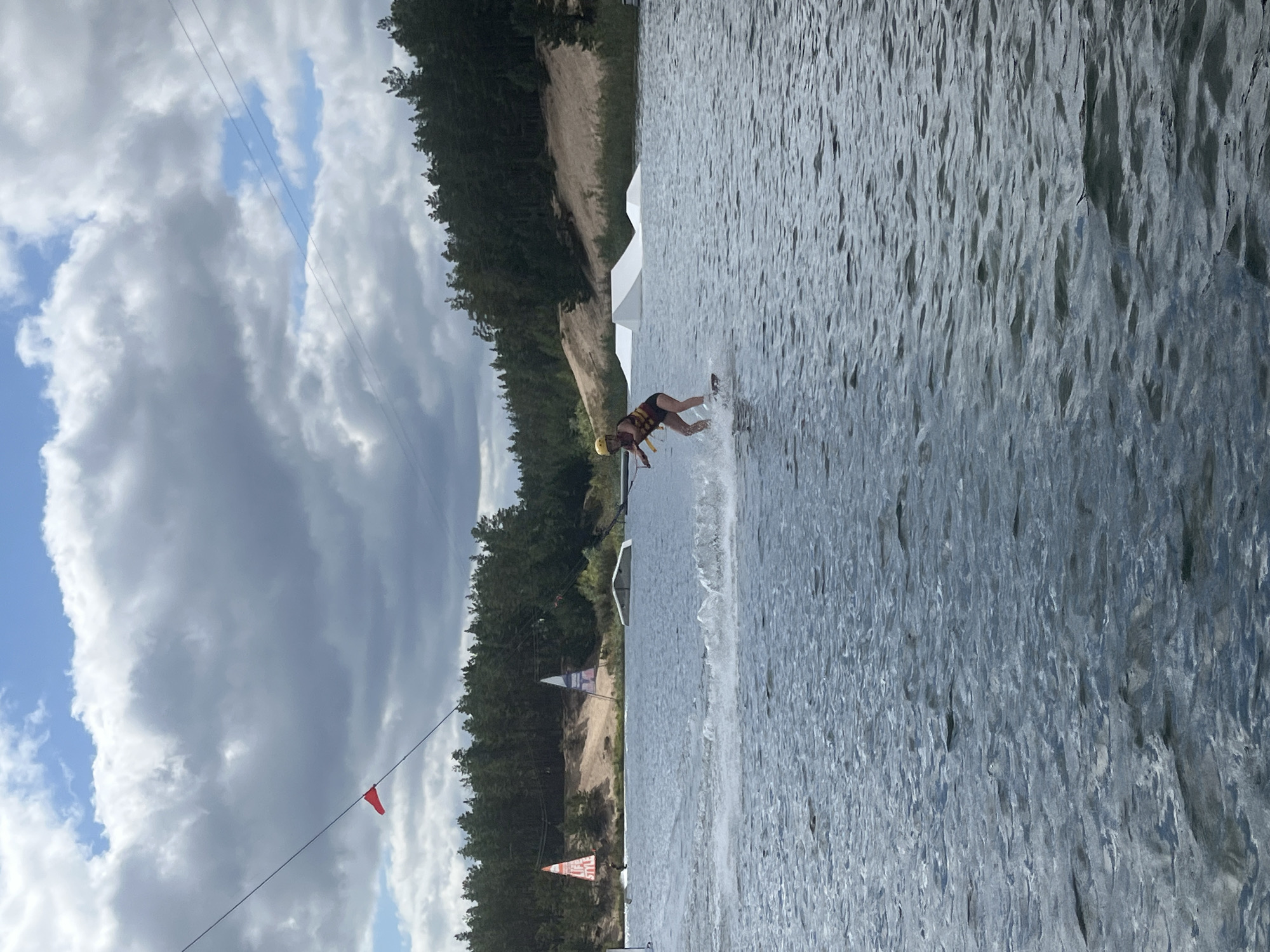 The image depicts a wakeboarding park in Männiku, Estonia. A lone individual is wakeboarding on a lake, creating a spray of water behind them. The wakeboarder is wearing a yellow helmet and a life vest. In the background, there are several white ramps designed for wakeboarding tricks. The lake is surrounded by a dense forest, and a sandy embankment is visible. The sky is partly cloudy with dramatic, grey clouds dominating the upper portion of the frame. Power lines are strung across the sky, and a small red flag is attached to one of them. Two large triangular banners are also visible near the shore, one with the text "LIFE STYLE" on it. The overall scene suggests an outdoor recreational activity taking place on a somewhat overcast day.