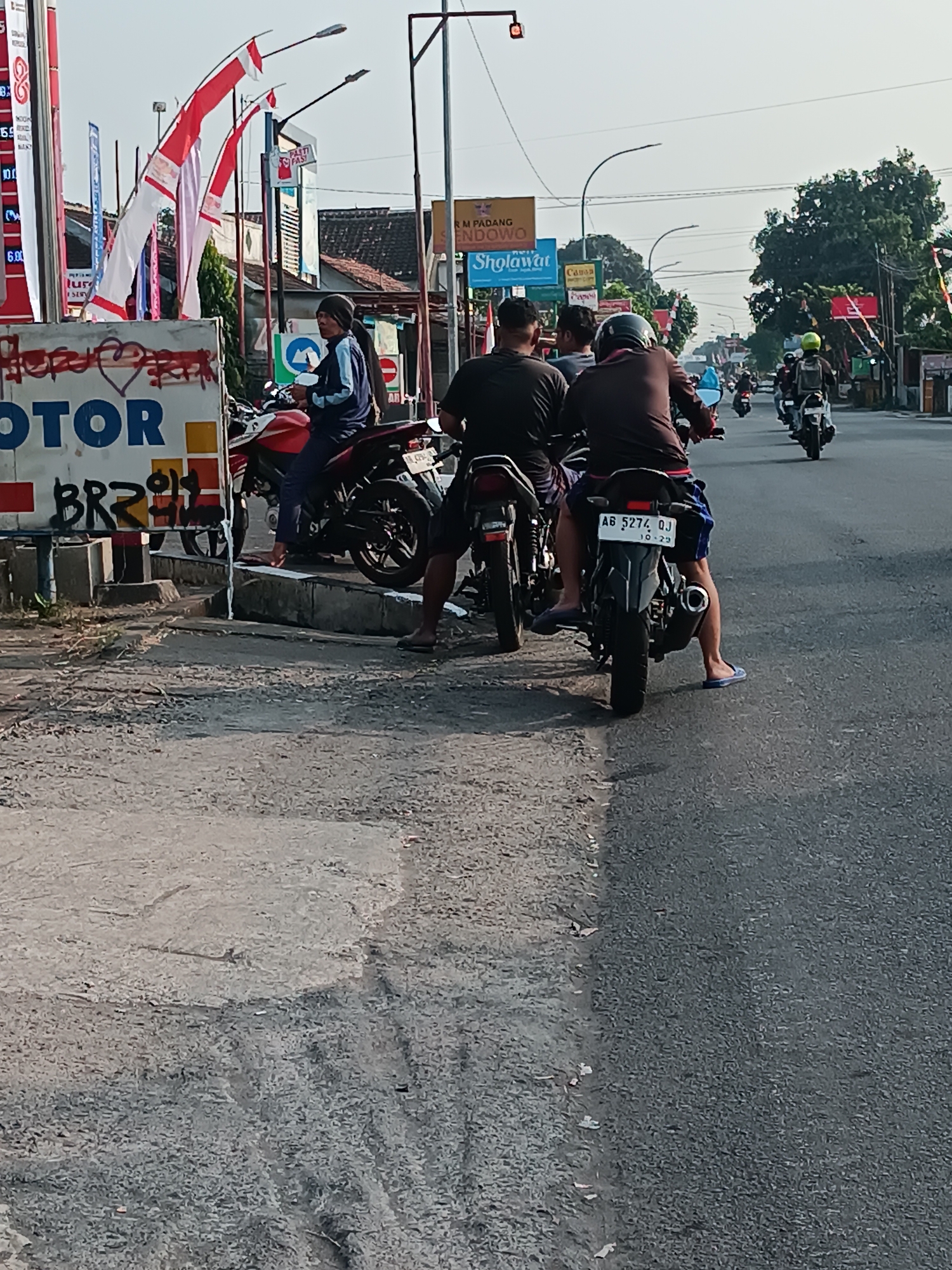 A street scene in Imogiri, Indonesia, features several people riding motorcycles. In the foreground, two men are seated on motorcycles, one facing away from the camera and the other partially visible. To the left, a woman in a blue jacket sits on a red motorcycle. A sign with "MOTOR" written on it is visible behind her. In the background, more motorcycles and people are on the road, with shops and trees lining the street. The sky is clear and bright.