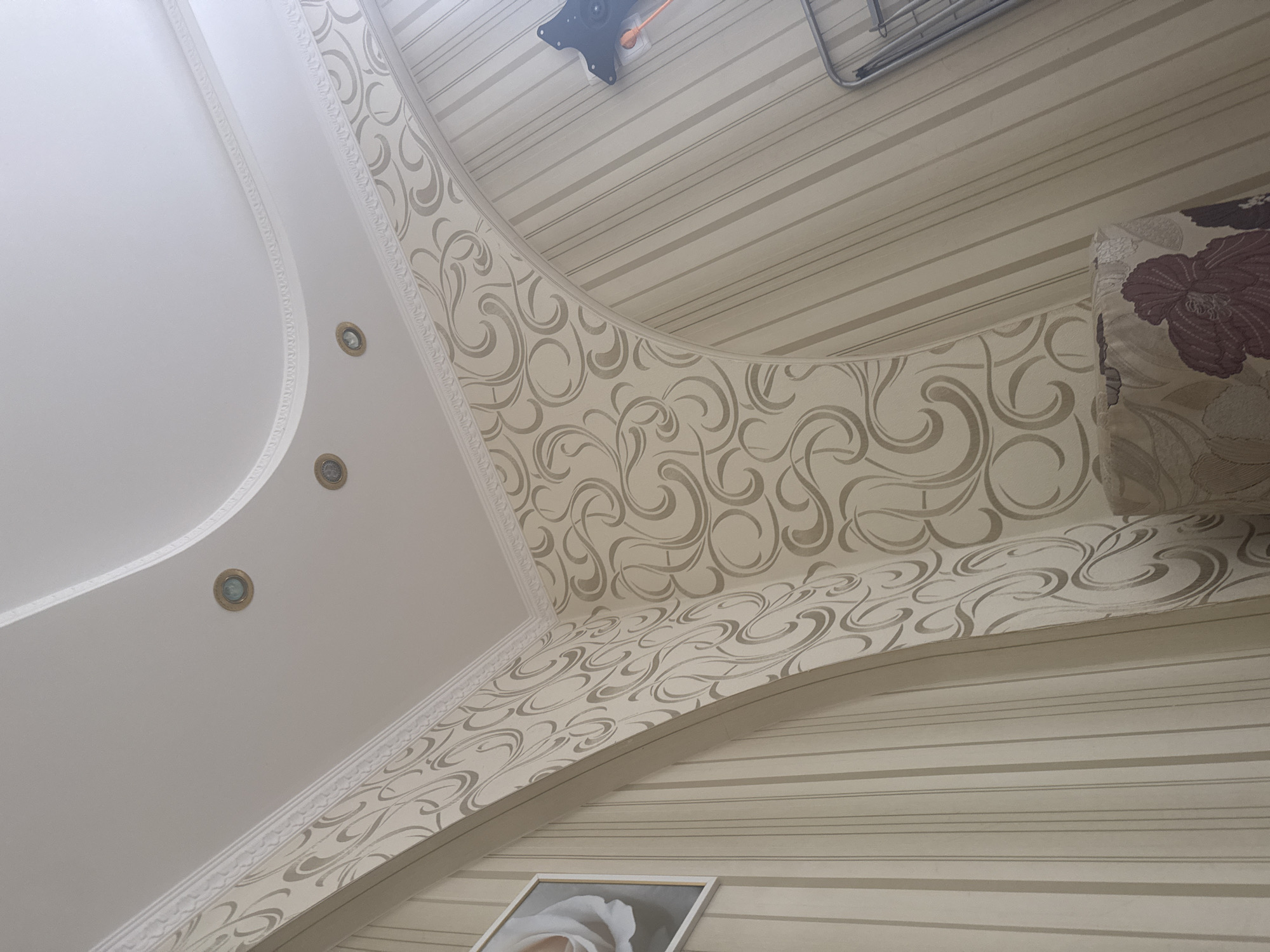 An interior shot looks up at a white ceiling with recessed lighting and decorative crown molding. The walls are covered in a beige wallpaper with a vertical stripe pattern and a swirling damask pattern in a lighter beige. There is a framed picture of a white rose on the left wall. A television mount is visible on the right wall. A patterned pillow or cushion is partially visible in the lower right corner. The image appears to be taken in a residential setting, possibly a living room or hallway.