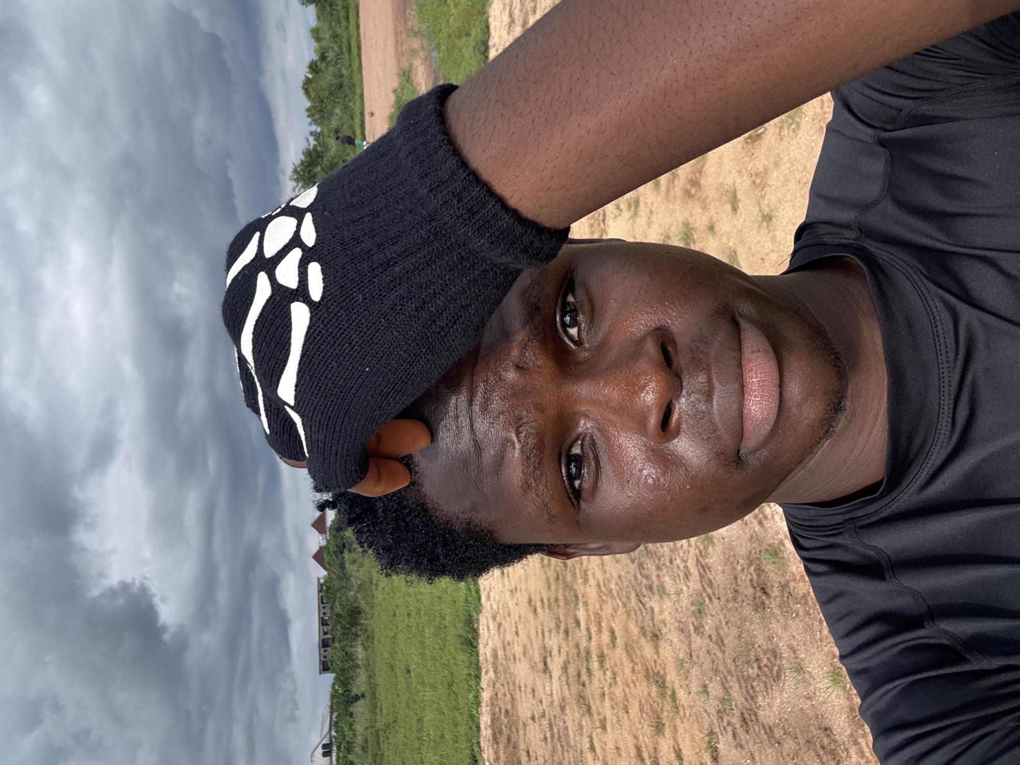 This media file features a close-up, low-angle shot of a young Black man outdoors. He is wearing a black long-sleeved shirt and a black glove with a white skeleton hand design on it, which he is using to touch his forehead. His skin appears to be glistening with sweat, suggesting physical exertion or warm weather. His gaze is directed towards the camera with a neutral expression.
The background is a mix of elements: to the left, a cloudy, overcast sky dominates, hinting at potentially stormy or at least humid weather. Further in the distance, there is greenery and what appear to be some partially constructed or abandoned buildings. To the right, the ground is a mix of dry, light brown earth and some patchy green vegetation.
The scene is set in Ihiagwa, Nigeria. The overall impression is one of an individual perhaps taking a break during an outdoor activity, possibly exercise, given the clothing and sweat. The lighting suggests it is daytime, but the cloudy sky creates a diffused and somewhat dramatic atmosphere. There is no visible text in the media file.