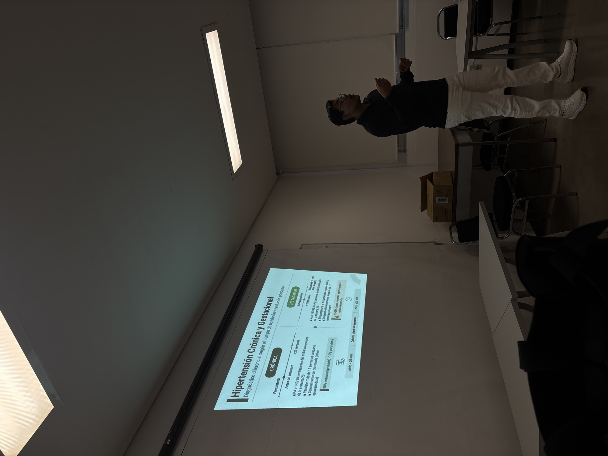 In a dimly lit classroom in Puebla, Mexico, a presentation is underway, projected onto a large screen. The screen displays a slide titled "Hipertensión Crónica y Gestacional," detailing differential diagnoses based on pregnancy and postpartum timelines. A man, dressed in a black long-sleeved shirt and white pants, stands to the right of the screen, facing it and gesturing with his hands as if explaining the content. He wears glasses. The classroom appears to be set up with tables and chairs, some of which are visible in the foreground and background. Boxes and other classroom materials are also present. The overall atmosphere suggests an academic lecture or presentation.