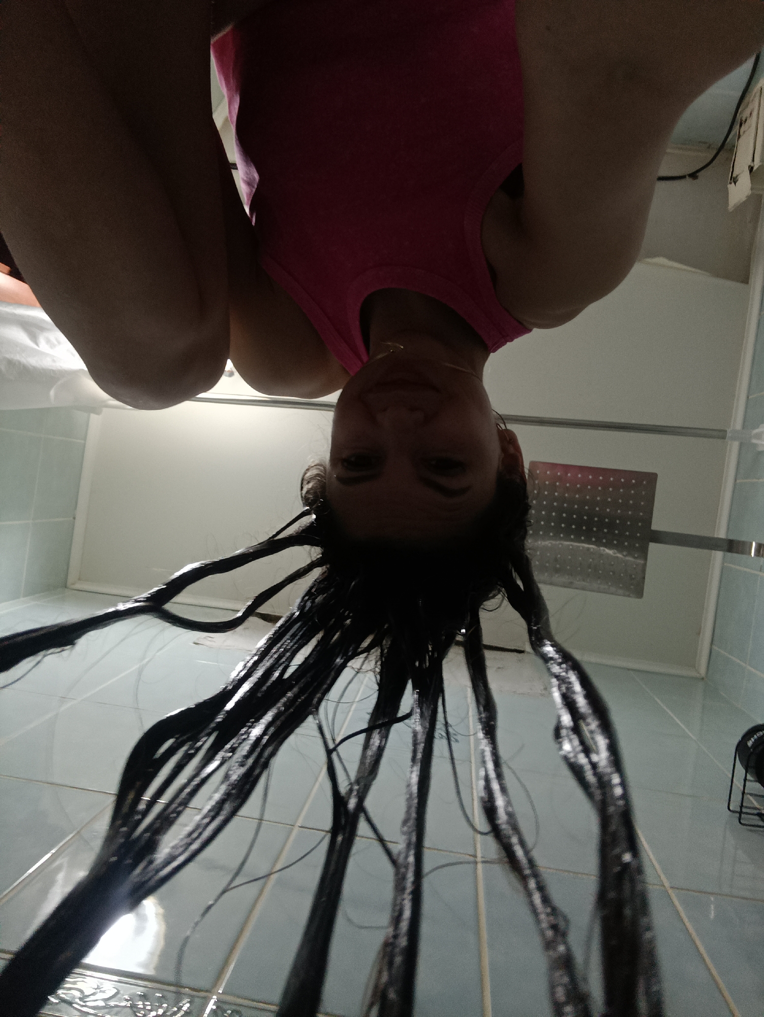 A woman in a pink tank top with black hair is upside down in a bathroom. The walls of the bathroom are blue tile, and there is a large shower head on the wall. Her hair is wet and stringy. The location is Unknown, Unknown.