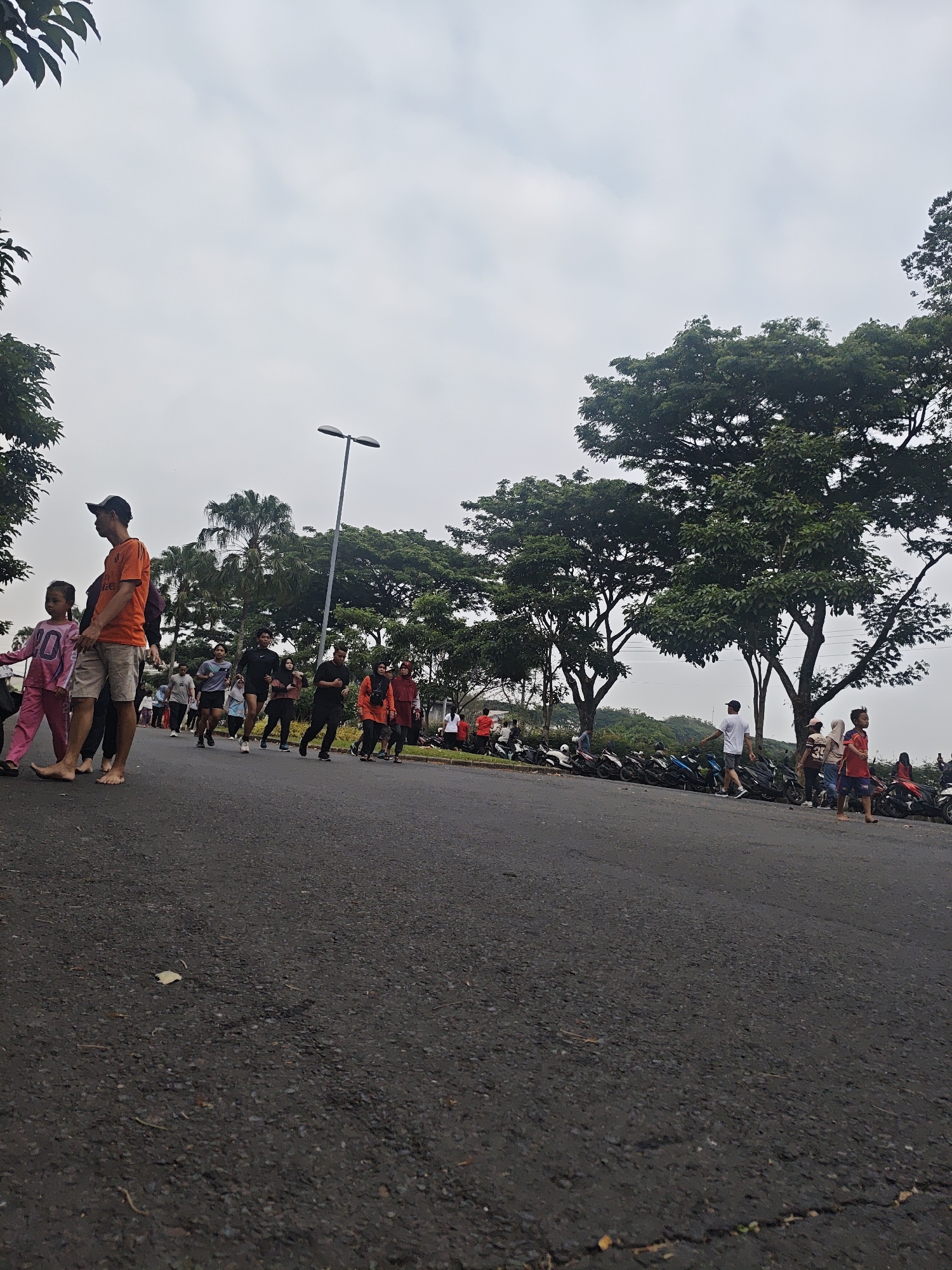 In Bacin, Indonesia, a group of people are participating in an outdoor event, possibly a race or a fun run. The event appears to be taking place on a paved road, with a lush green landscape of trees and foliage in the background. The sky is overcast, suggesting a cloudy day.
In the foreground, on the left side of the road, a young girl in a pink outfit is walking barefoot. Beside her, a man in an orange t-shirt and light-colored shorts is looking towards the left. Behind them, another young girl in a pink outfit with the number "20" on it is also walking.
Further down the road, a group of adults are seen running. They are dressed in various athletic wear, including t-shirts, shorts, and leggings. Some are wearing athletic shoes, while others appear to be running barefoot.
In the middle ground, a street lamp stands tall on the right side of the road. The background is dominated by mature trees with dense green canopies. Beyond the trees, more foliage and a hint of a hilly terrain can be seen.
To the right of the road, a line of parked motorcycles is visible. Several people are standing or walking near the motorcycles, some of whom are also observing the event. A few individuals, including a person in a red shirt and another in a white t-shirt, are more clearly visible among the spectators.
The overall scene depicts a community gathering focused on physical activity in a natural outdoor setting.