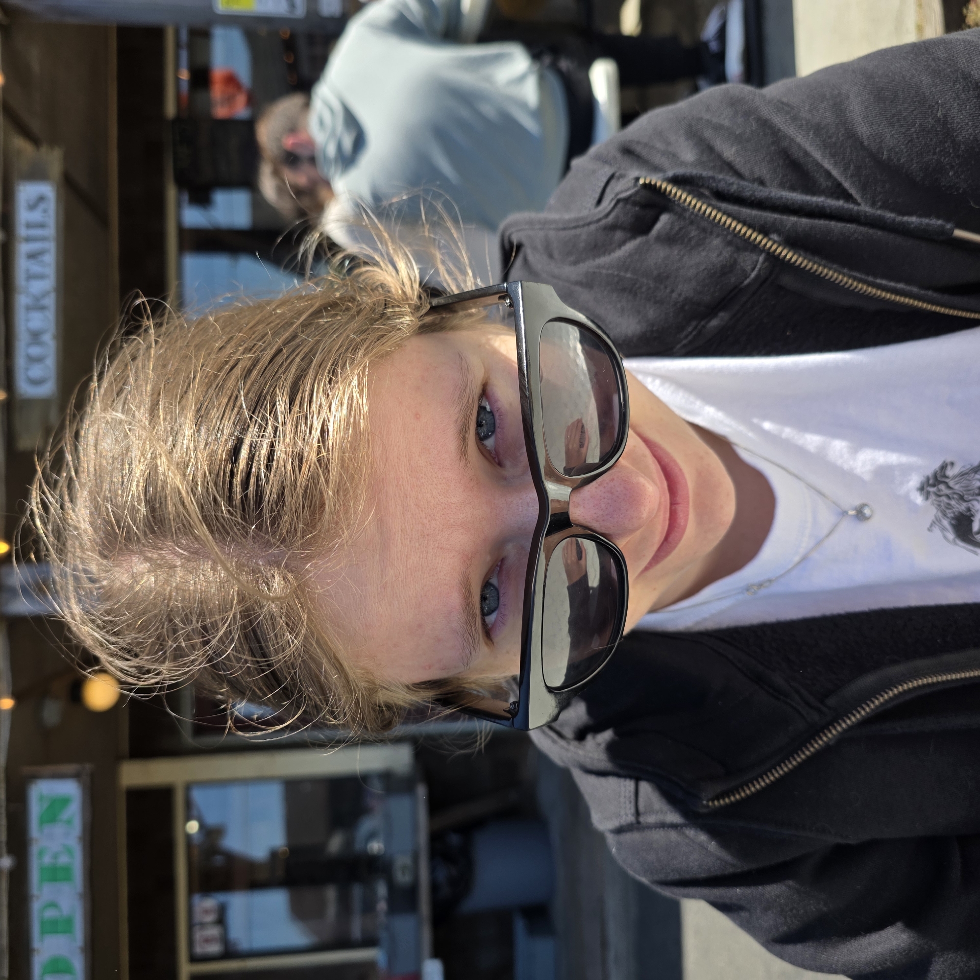A headshot of a young person with blonde hair and blue eyes is centered in the frame, looking down at the camera with a slight smile. They are wearing black sunglasses and a white t-shirt with a silver necklace. A black zip-up hoodie is visible around their shoulders. In the background, there is a building with a sign that reads "OPEN" in green letters and another sign that says "COCKTAILS". Other people are blurred in the background. The scene appears to be outdoors, possibly a restaurant or bar, in Cannon Beach, Oregon, United States of America, indicated by the architecture and the style of the signage.