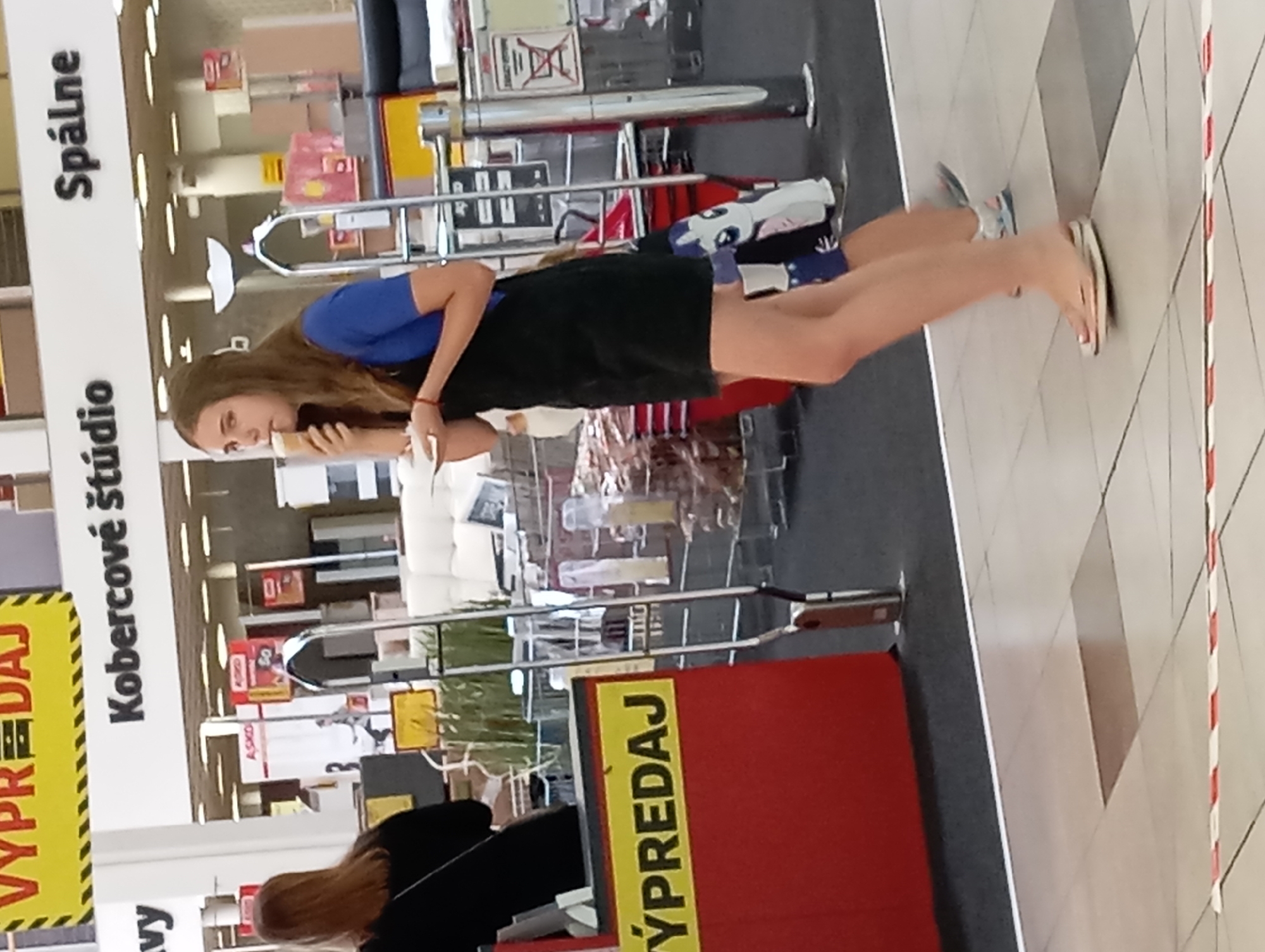 The image captures an indoor scene, likely a retail environment, given the signage visible. In the foreground, a young woman with long brown hair is walking and eating an ice cream cone. She is wearing a blue short-sleeved shirt and a black overall dress, with flip-flops on her feet. Behind her, another person, possibly a child, is partially visible wearing shorts and sneakers.
The background features large signage, including "VYPREDAJ" (SALE in Slovak) in yellow and red, and "Kobercové štúdio" (Carpet Studio) and "Spálne" (Bedrooms) in black letters on a white background. These signs suggest the location is likely a department store or a large furniture and home goods retailer. The environment includes display shelves, racks, and what appear to be furniture items, such as sofas. The flooring is tiled. The overall impression is a busy commercial space. While the image does not explicitly show outdoor elements, the context of such a large retail establishment suggests it would be located within the city of Nitra, Slovakia, given the Slovak language on the signage.