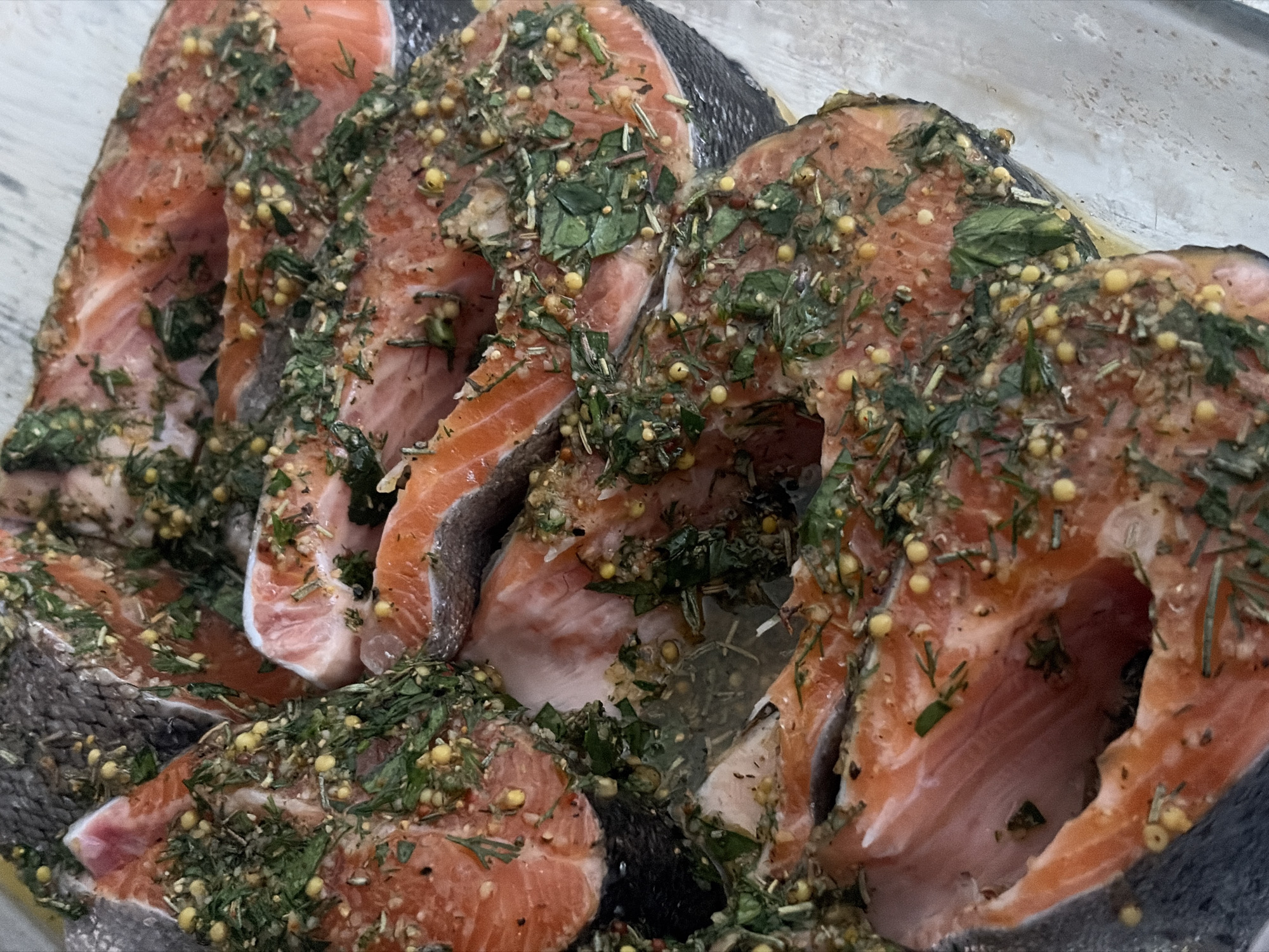 The image shows several cuts of salmon, possibly steaks, marinated with green herbs and yellow seeds, arranged in a clear container. The salmon pieces are orange-pink with visible skin on some cuts. The herbs appear to include dill and basil, and the seeds are likely mustard seeds. The container sits on a light-colored, possibly wooden, surface. The scene doesn't provide visual cues related to the city of Saint Petersburg, Russia.
