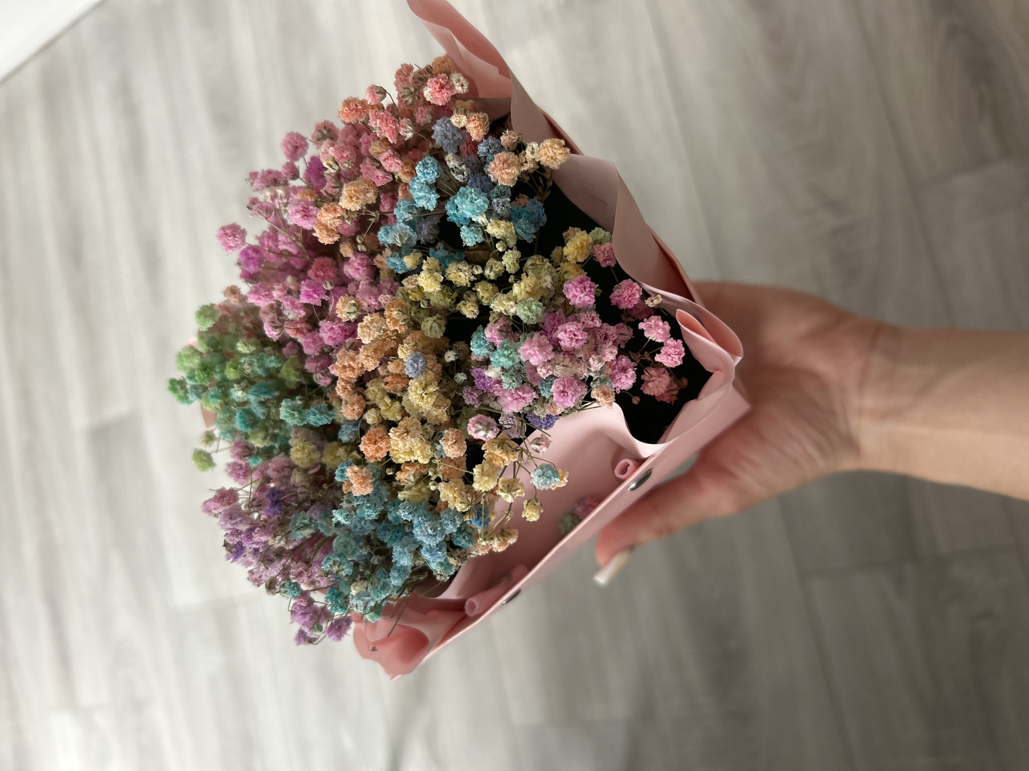 A hand is holding a bouquet of dyed baby's breath flowers in various pastel colors. The flowers are small and clustered together, creating a fluffy appearance. The bouquet is wrapped in a pink paper. The background is a light-colored wooden floor.