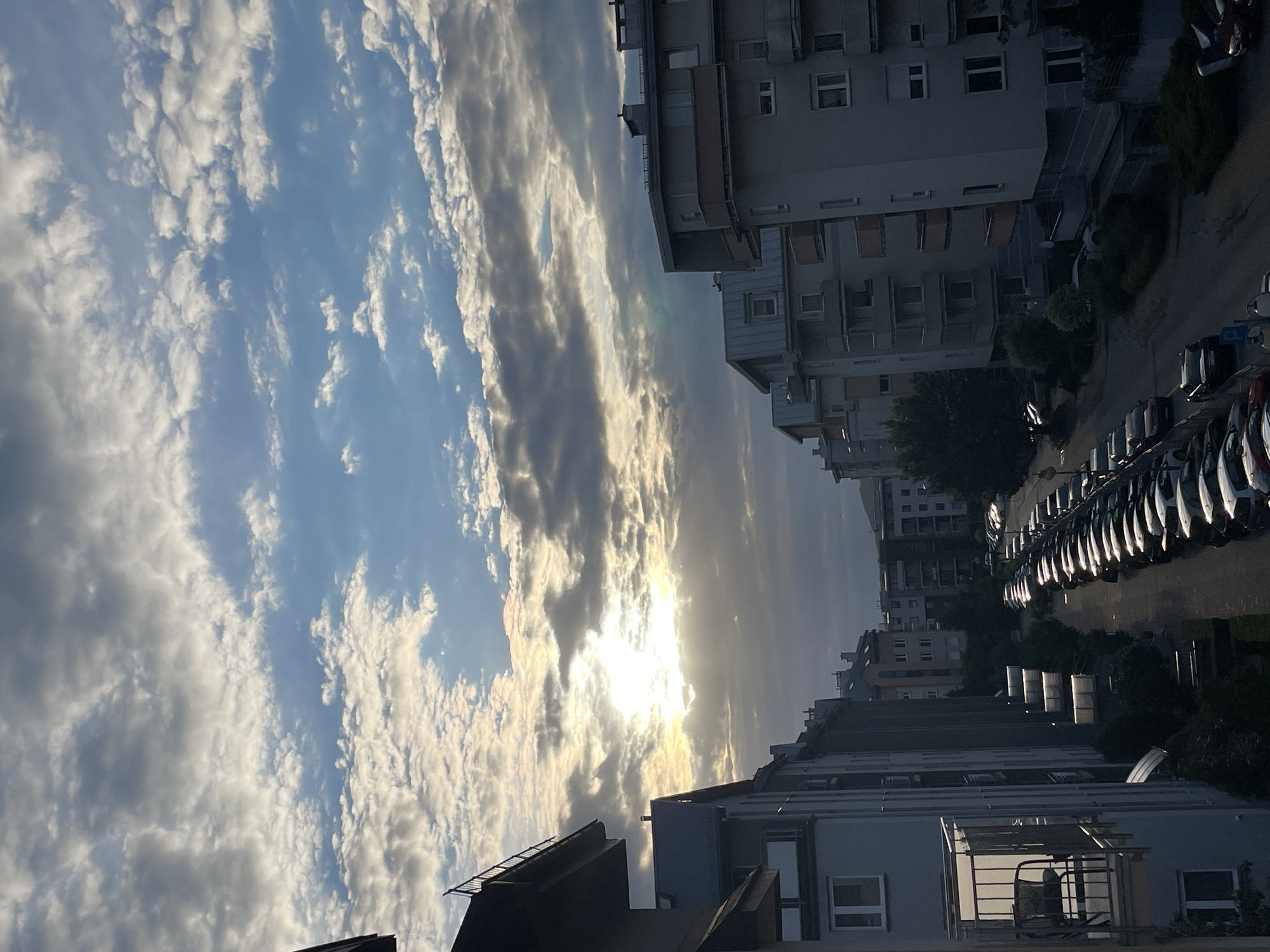 The image shows a street lined with apartment buildings and parked cars, viewed from an elevated perspective. The sun is partially obscured by clouds, casting a warm glow and rays of light through the sky. The buildings are modern in design, with balconies and windows visible. A row of cars is parked along the side of the street, their surfaces reflecting the light. The sky is a mix of blue and gray clouds, with some areas of bright sunlight. The overall scene suggests a residential area in a city.