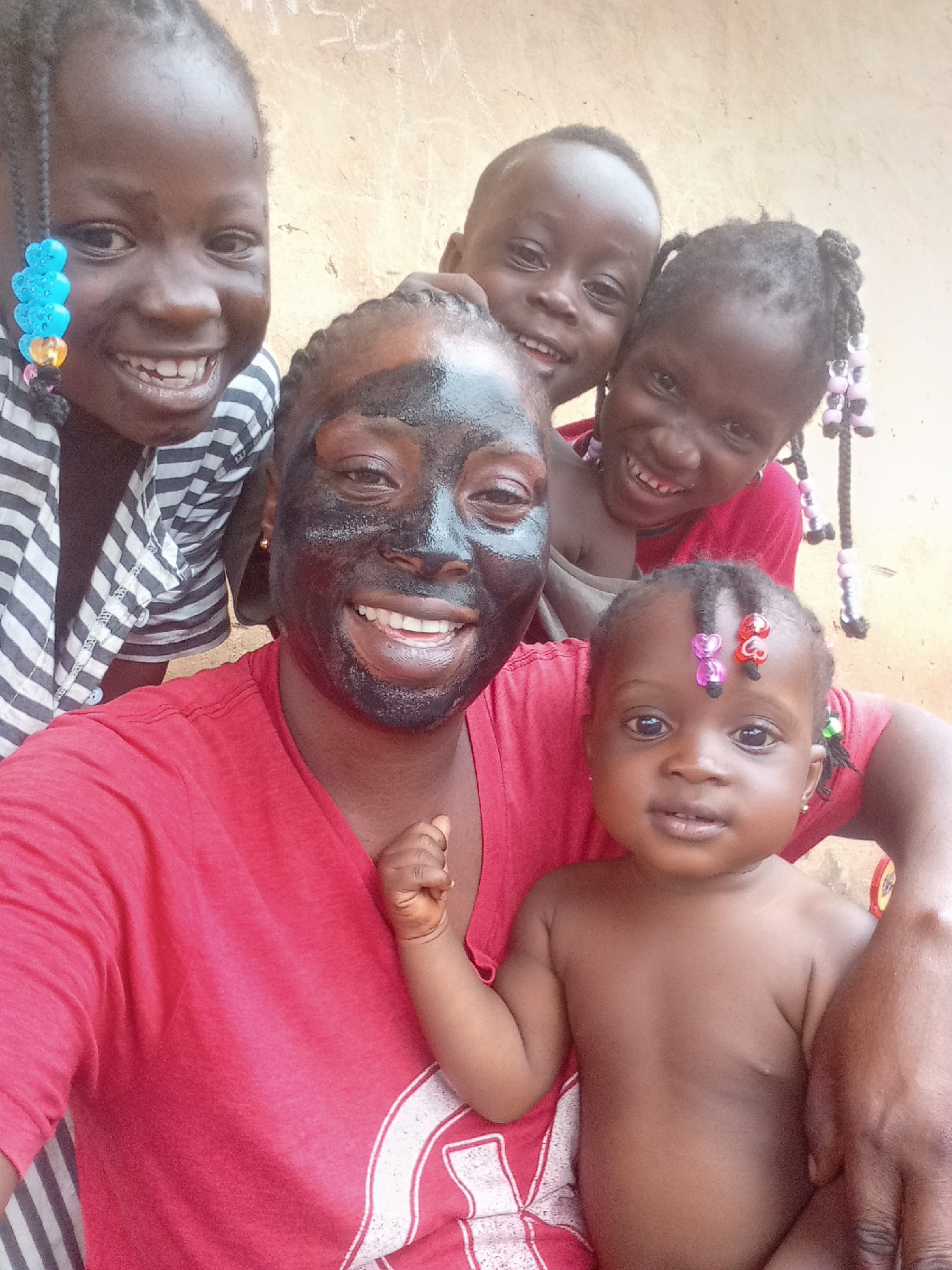 A selfie shows a woman wearing a black face mask smiling widely. Four children are clustered around her. The children have braided hair, and some braids are decorated with beads. The woman wears a red t-shirt. The child in the front is unclothed and appears to be an infant. Behind the woman are the smiling faces of three children. A cream-colored wall is in the background. The photograph was taken in Akwanga, Nigeria.