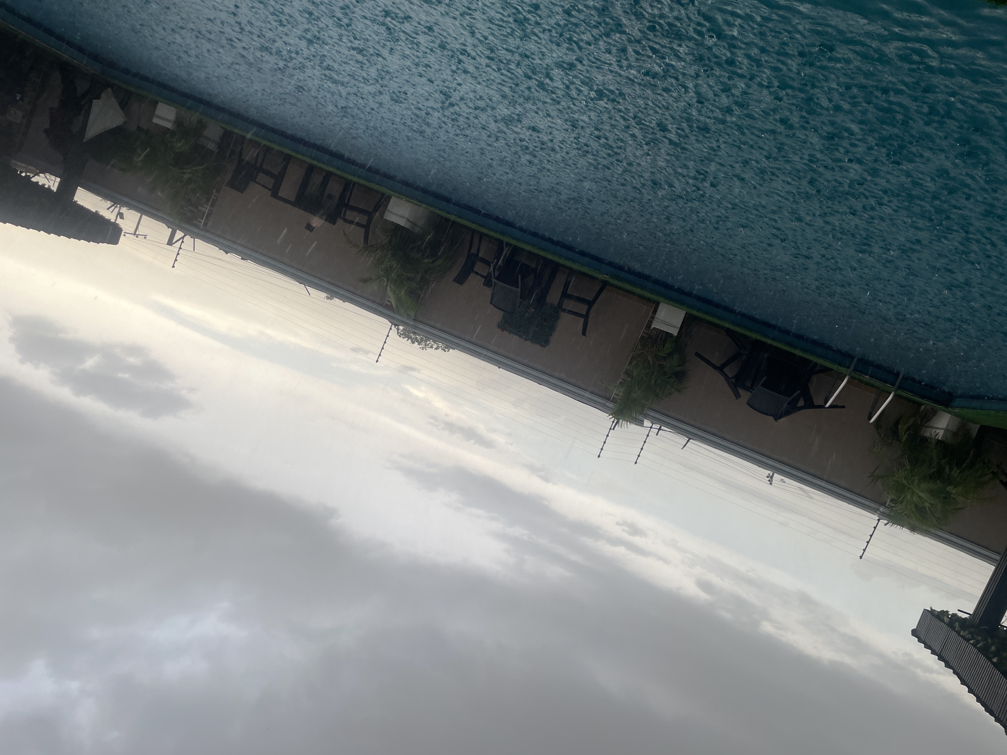 This image captures an outdoor scene featuring a swimming pool, a patio area, and an overcast sky, taken during a rain shower.
The upper half of the frame is dominated by a cloudy sky, displaying various shades of gray and white, indicating heavy cloud cover. Below the sky, a patio or outdoor deck is visible, furnished with several dark, outdoor chairs and possibly tables, positioned against a light-colored building wall or structure. Lush green foliage, including what appear to be palm trees and potted plants, can be seen along this wall. A series of parallel wires, resembling an electric fence, runs horizontally across the mid-ground, separating the sky from the patio area. On the far right edge of the frame, a dark, angled structure, possibly part of a building's roof, is partially visible.
The lower half of the image features a swimming pool with turquoise water. The surface of the pool is visibly disturbed by numerous raindrops, creating extensive ripples and splashes, clearly indicating that it is raining. No people are present in the scene. A small, white sign with unreadable text is visible on a post or wall near the pool. The weather is overcast and rainy, creating a diffused light during what appears to be daytime. The setting is an outdoor recreational area, possibly a hotel or resort, in a tropical or sub-tropical region like Abuja, Nigeria. The image conveys a quiet, atmospheric moment during the rain.