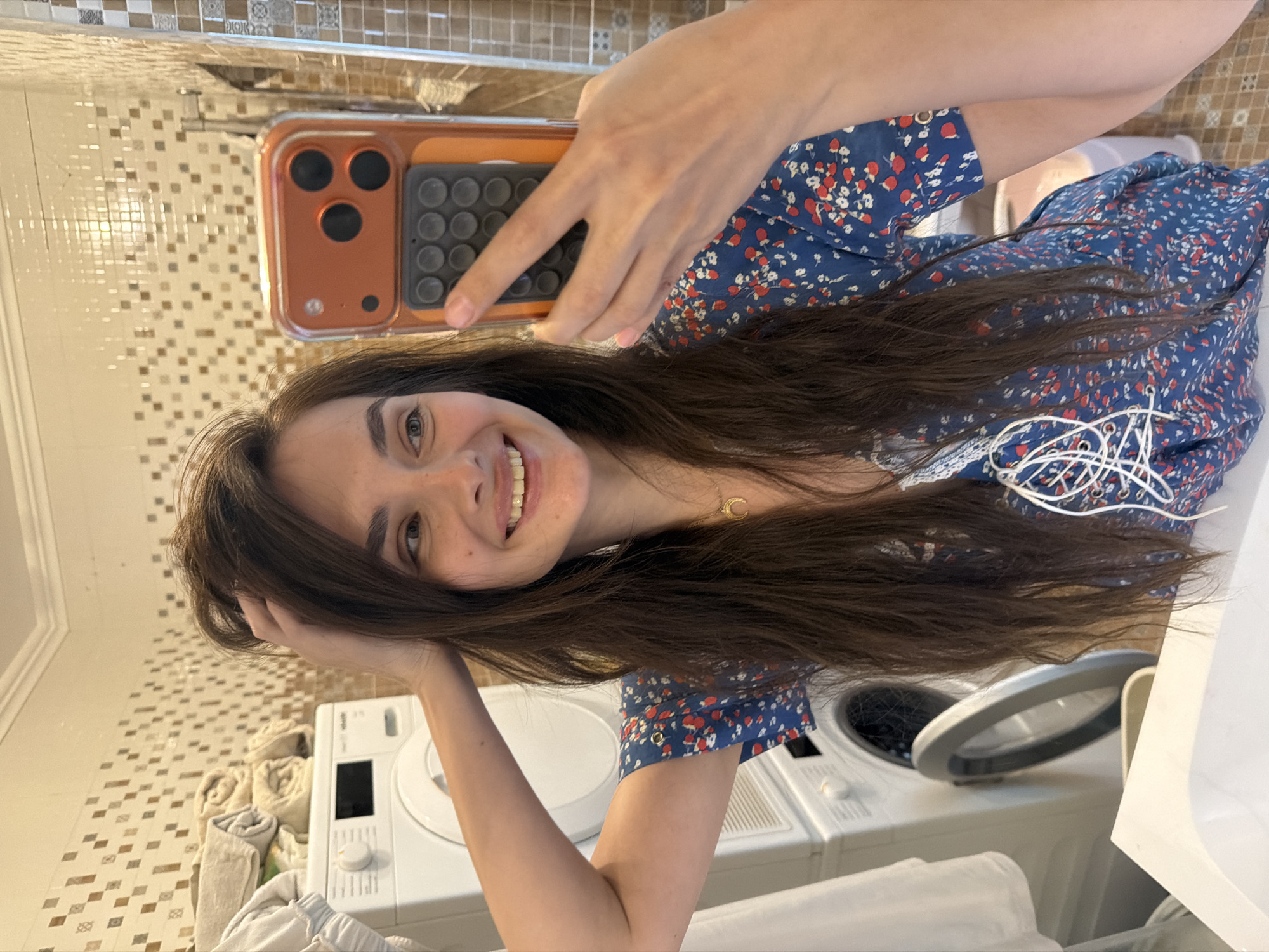 The image shows a young woman taking a selfie in a bathroom. She is wearing a blue floral dress. Her hair is long and dark brown, and she is smiling. Her right hand is holding her phone, and her left hand is resting on her head. In the background, there is a washing machine and a tiled wall. The time of day is not discernible, and there are no indications of weather conditions. The location is Ufa, Russia. The overall mood of the image is casual and relaxed. No visible text is present in the image.