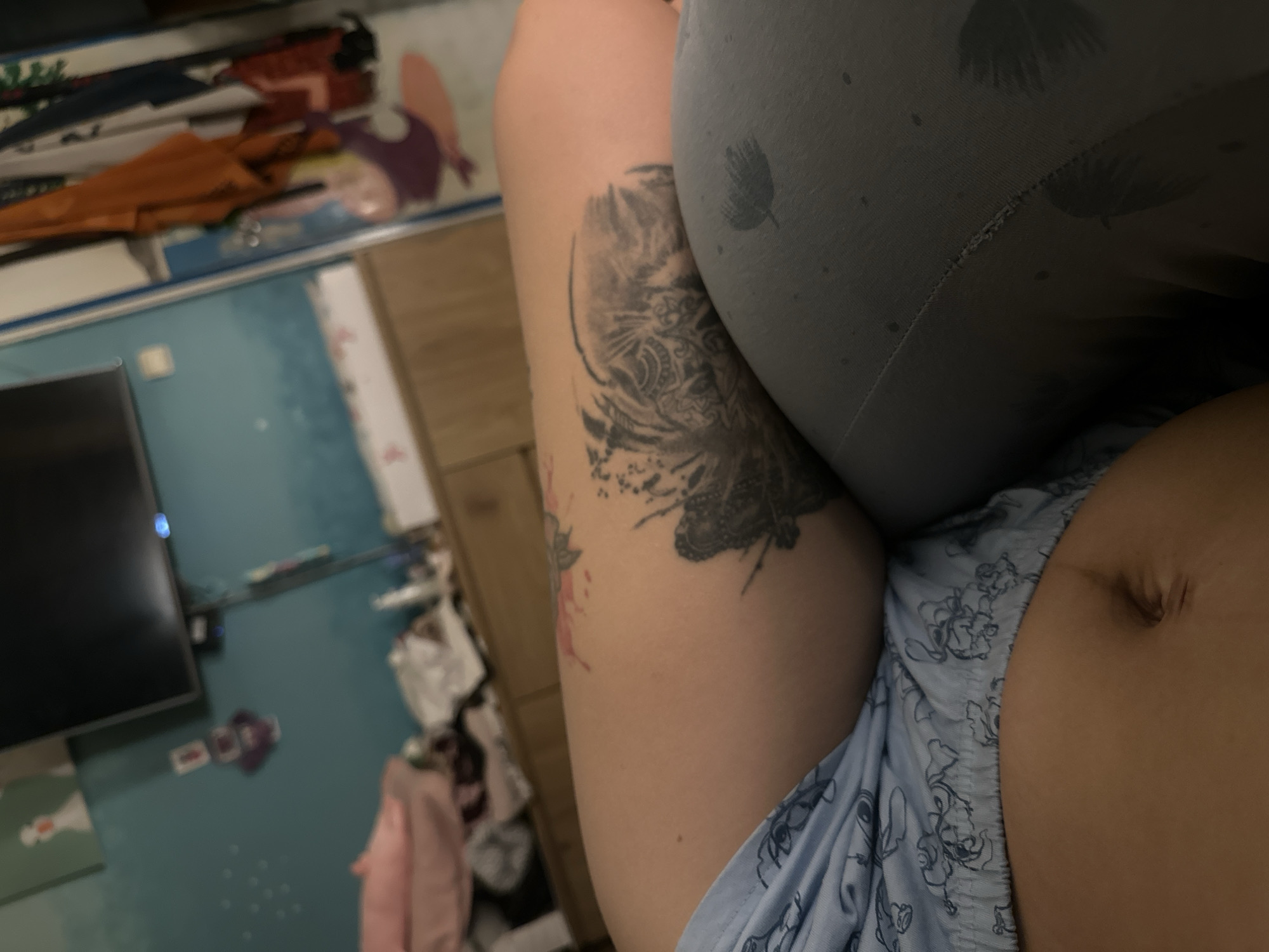 The image shows a close-up view of a person's abdomen and thigh, with a tattoo of a tiger's face on the thigh. The person is lying down, wearing blue patterned pajamas on their lower body and gray leggings on their upper body. The background is blurred, but it appears to be a room with a television on the wall, a dresser, and some clothing hanging on a rack. The setting is likely a bedroom. The location in Plovdiv, Bulgaria cannot be determined from the image.